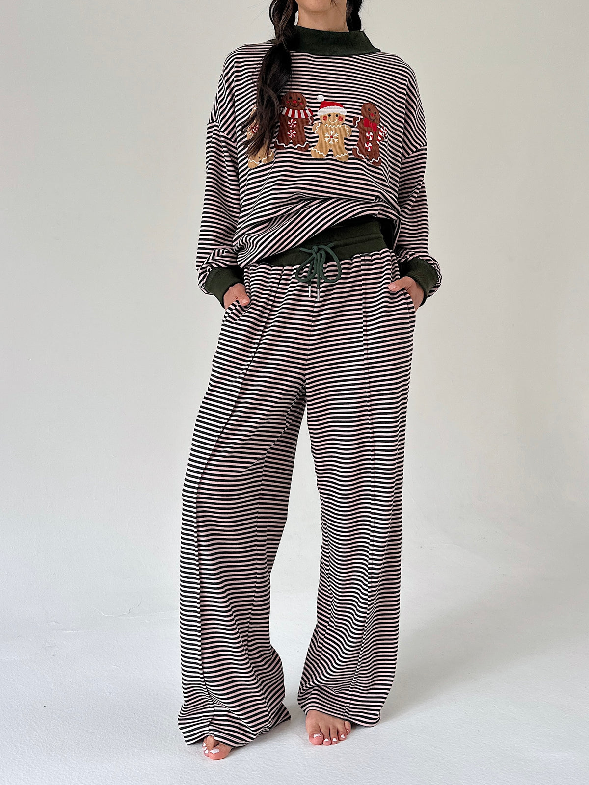 A woman modeling a matching holiday-themed loungewear set consisting of a long-sleeve top and wide-leg pants, both featuring a horizontal stripe pattern with a festive gingerbread cookie graphic on the chest.