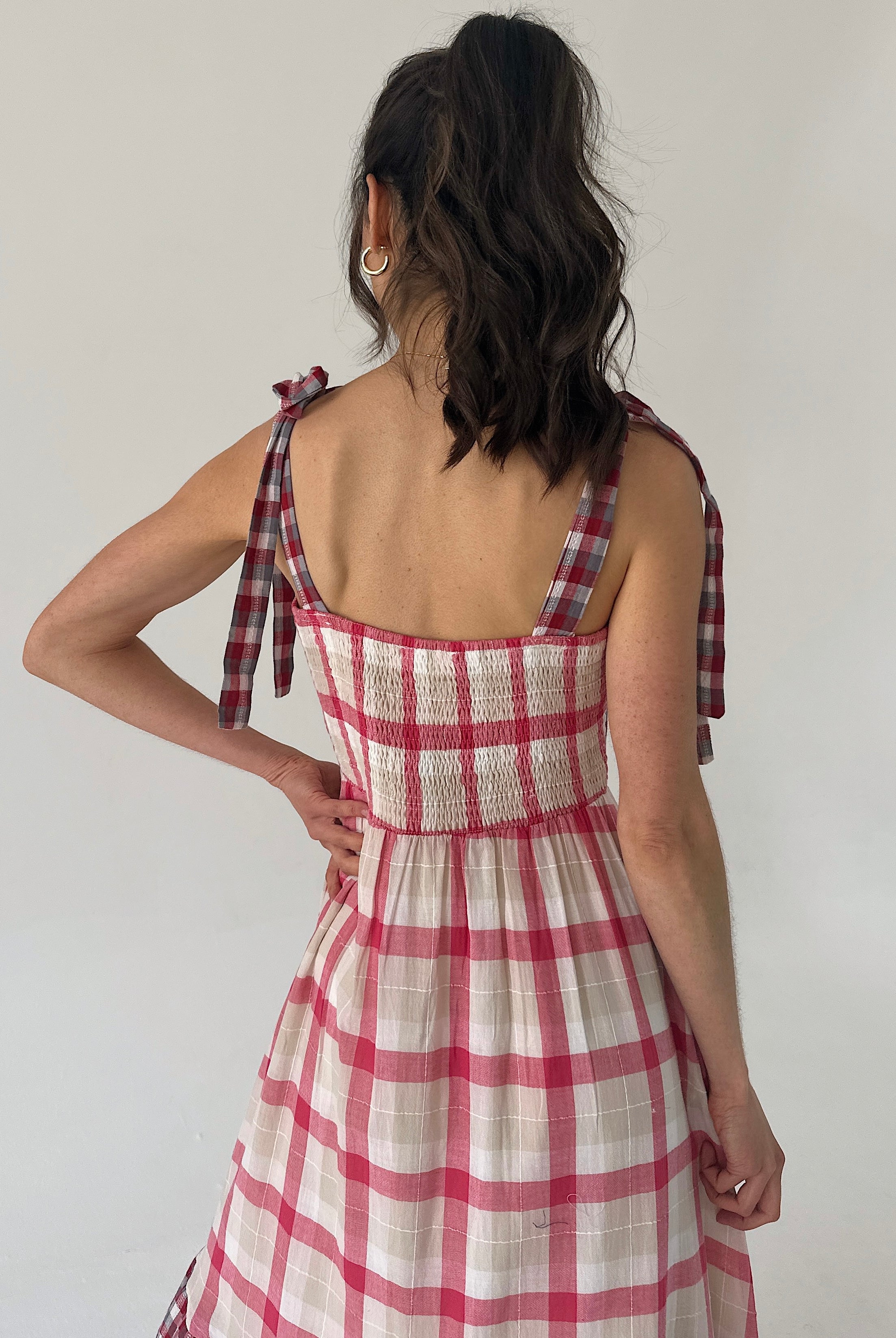 Woman wearing a red and white checkered dress with bow straps and smocked back on a plain background