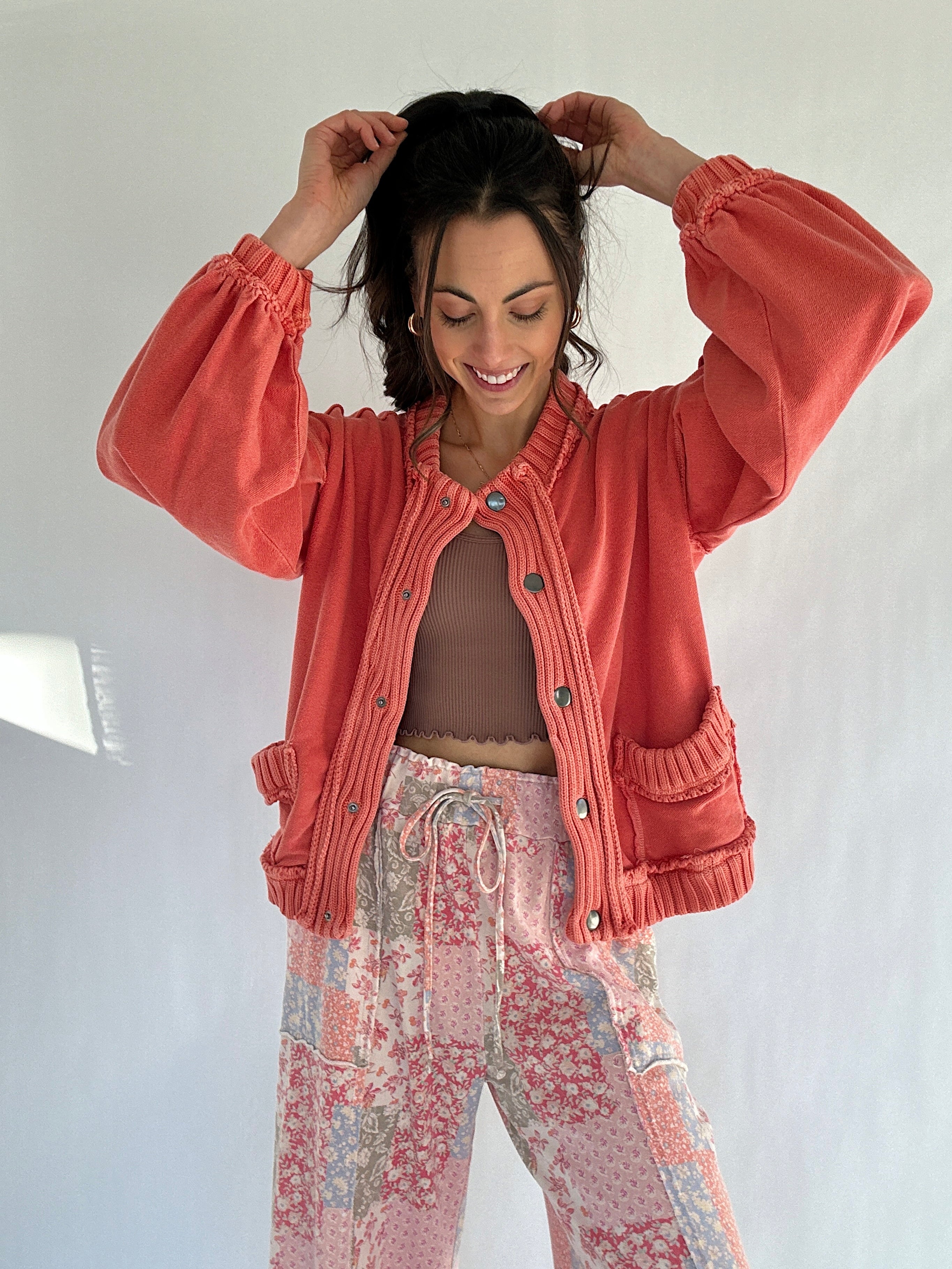 Woman wearing a coral jacket and floral pants against a white background putting her hair up