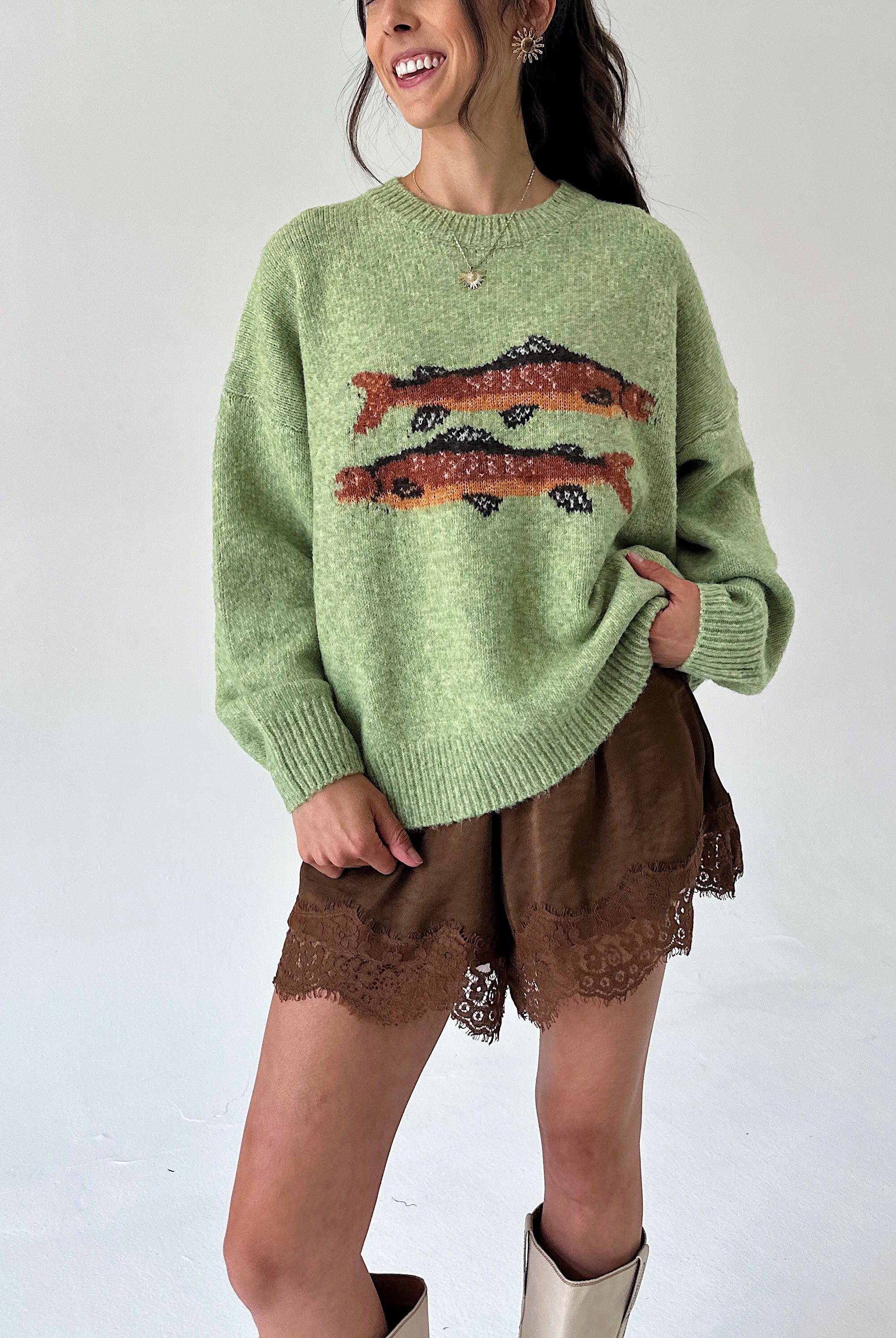 A woman wearing a light green knit sweater with two orange and black fish motifs across the chest, paired with brown lace-trim shorts and beige boots, against a plain white background.