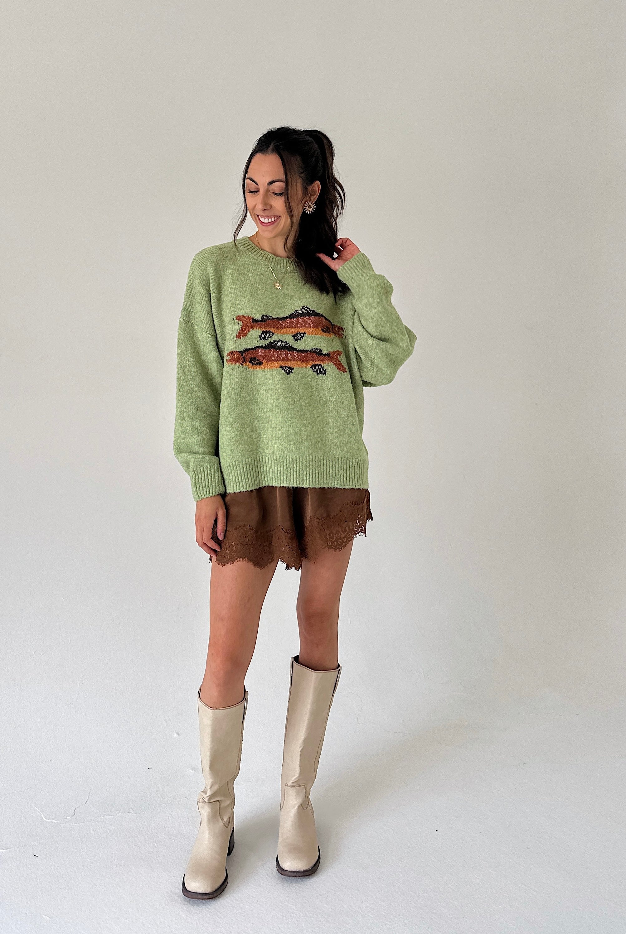 A woman modeling a light sage green knit sweater with a colorful fish graphic across the chest, paired with a brown satin shorts and beige knee-high boots, against a plain light gray studio background.