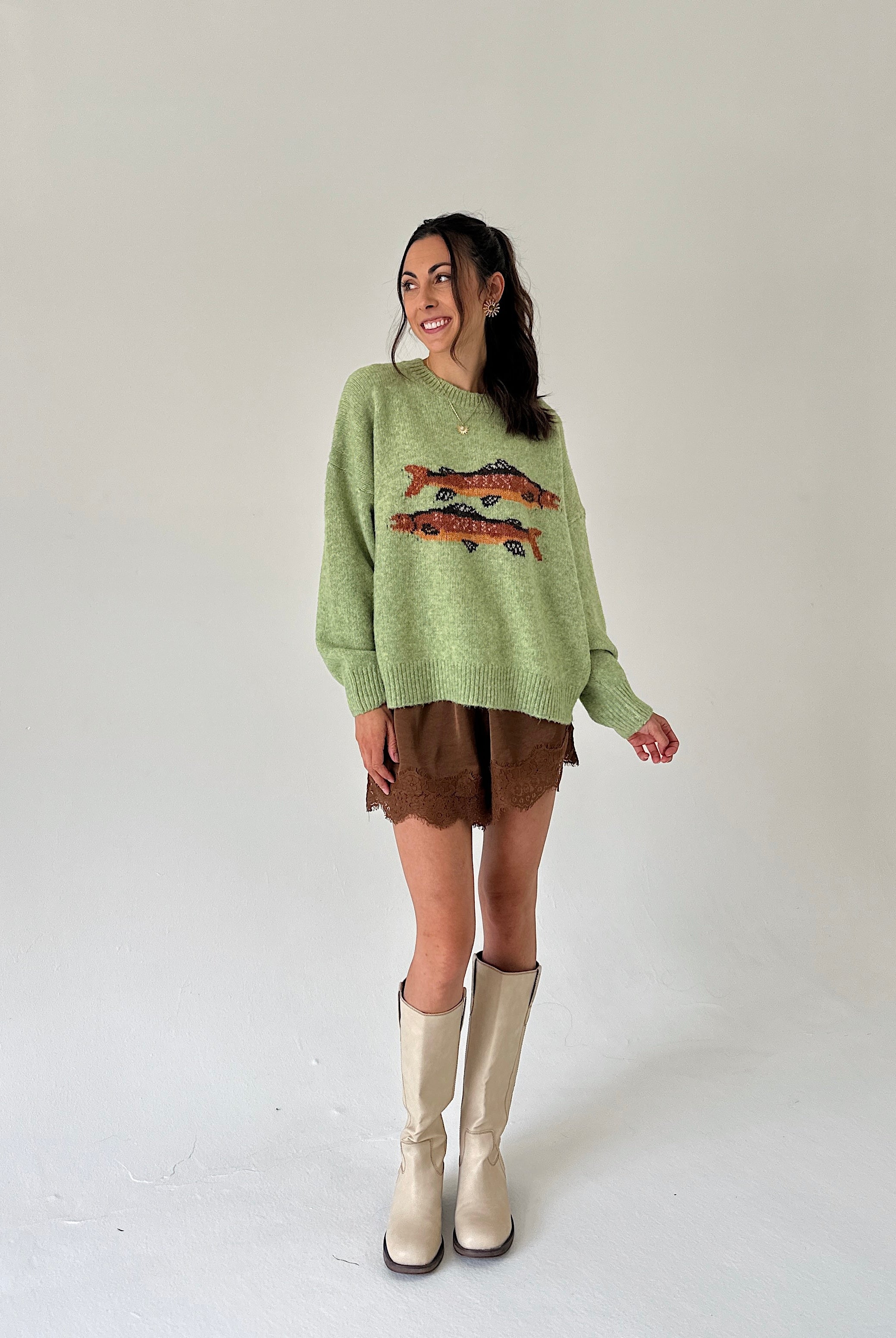 A woman modeling a light green knit sweater with a fish graphic, paired with brown shorts and beige knee-high boots, against a plain light gray studio background.