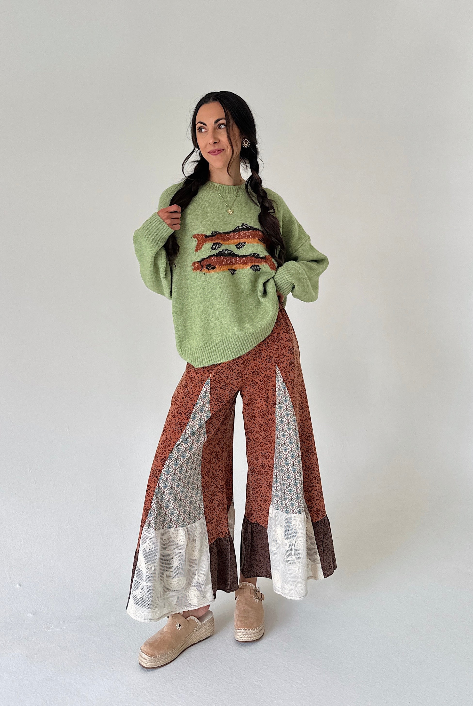 A woman modeling a light green fuzzy knit sweater with a colorful trout graphic, paired with rust-brown wide-leg patterned pants featuring lace accents and beige slip-on shoes, against a plain light gray studio background.