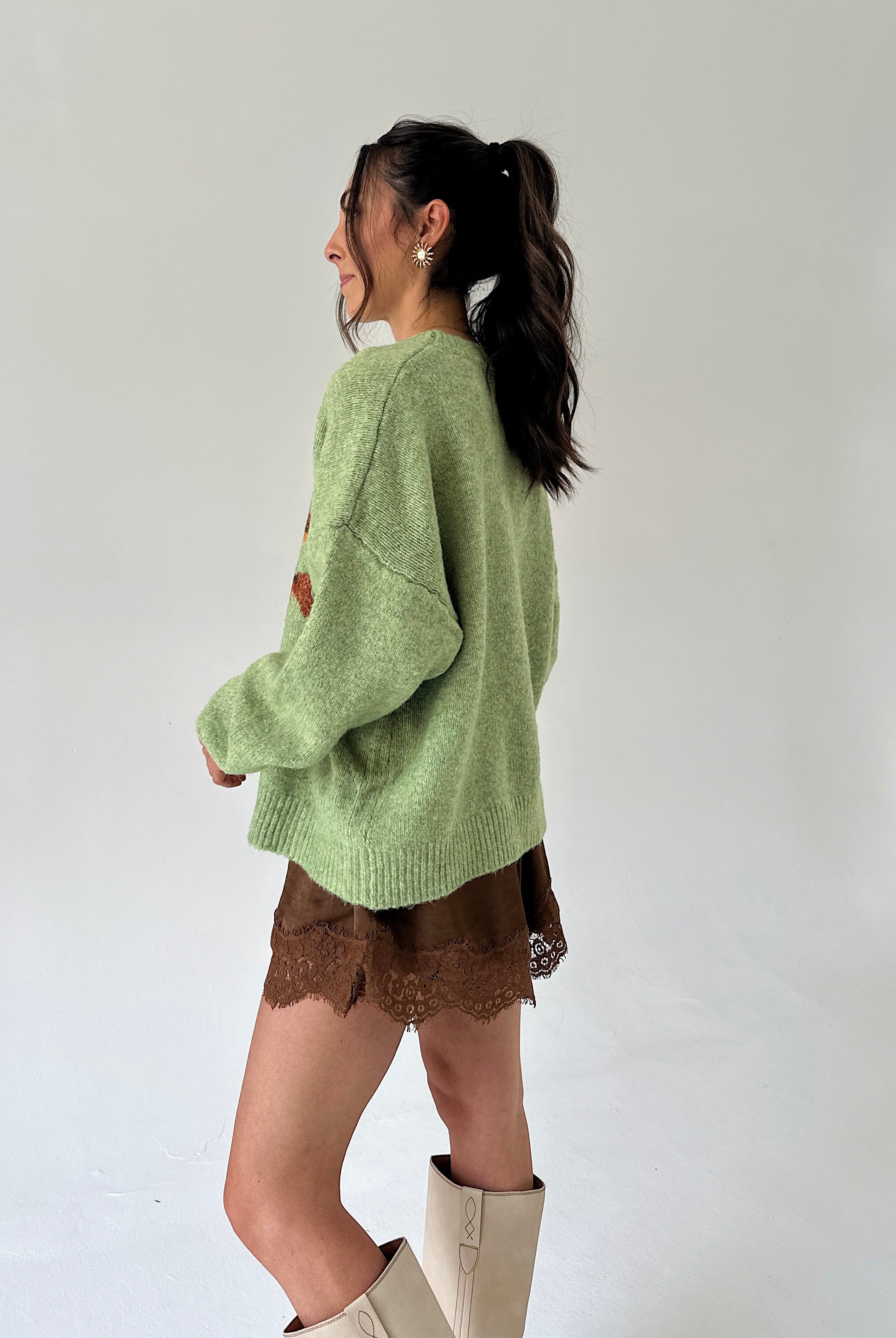 A woman wearing a light sage green knit sweater, brown lace shorts, and beige boots, photographed from the side against a plain white studio background.