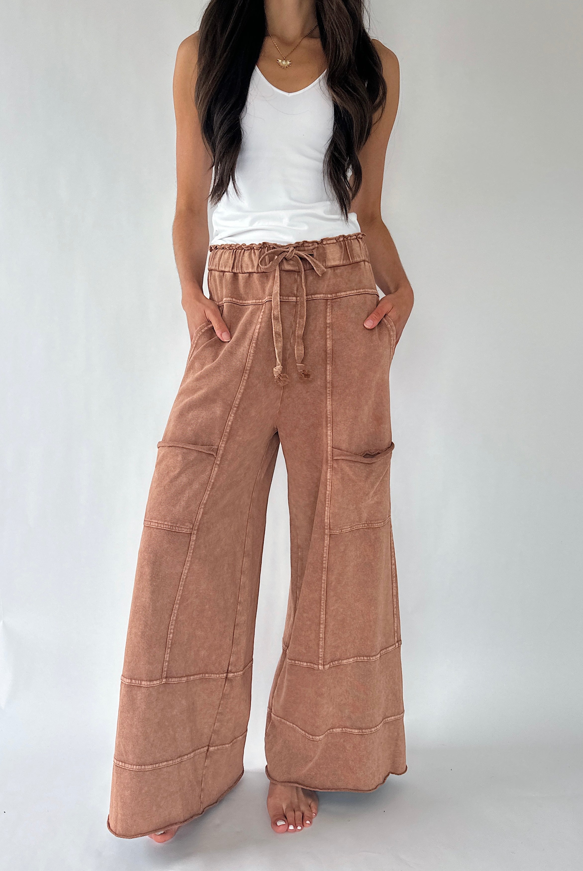 A woman wearing wide-leg, high-waisted pants in a dusty rose-brown with a drawstring waist and front patch pockets, paired with a white tank top, standing against a light gray studio background.