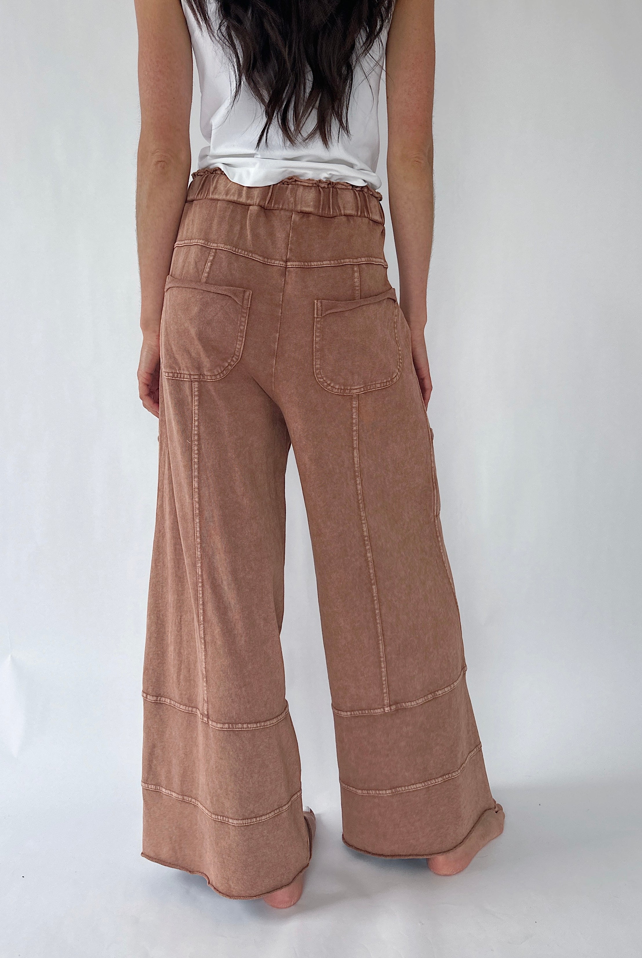 A person shown from the back wearing high-waisted, wide-leg brown pants with a ruffled waistband, a white sleeveless top, and bare feet against a plain light background.