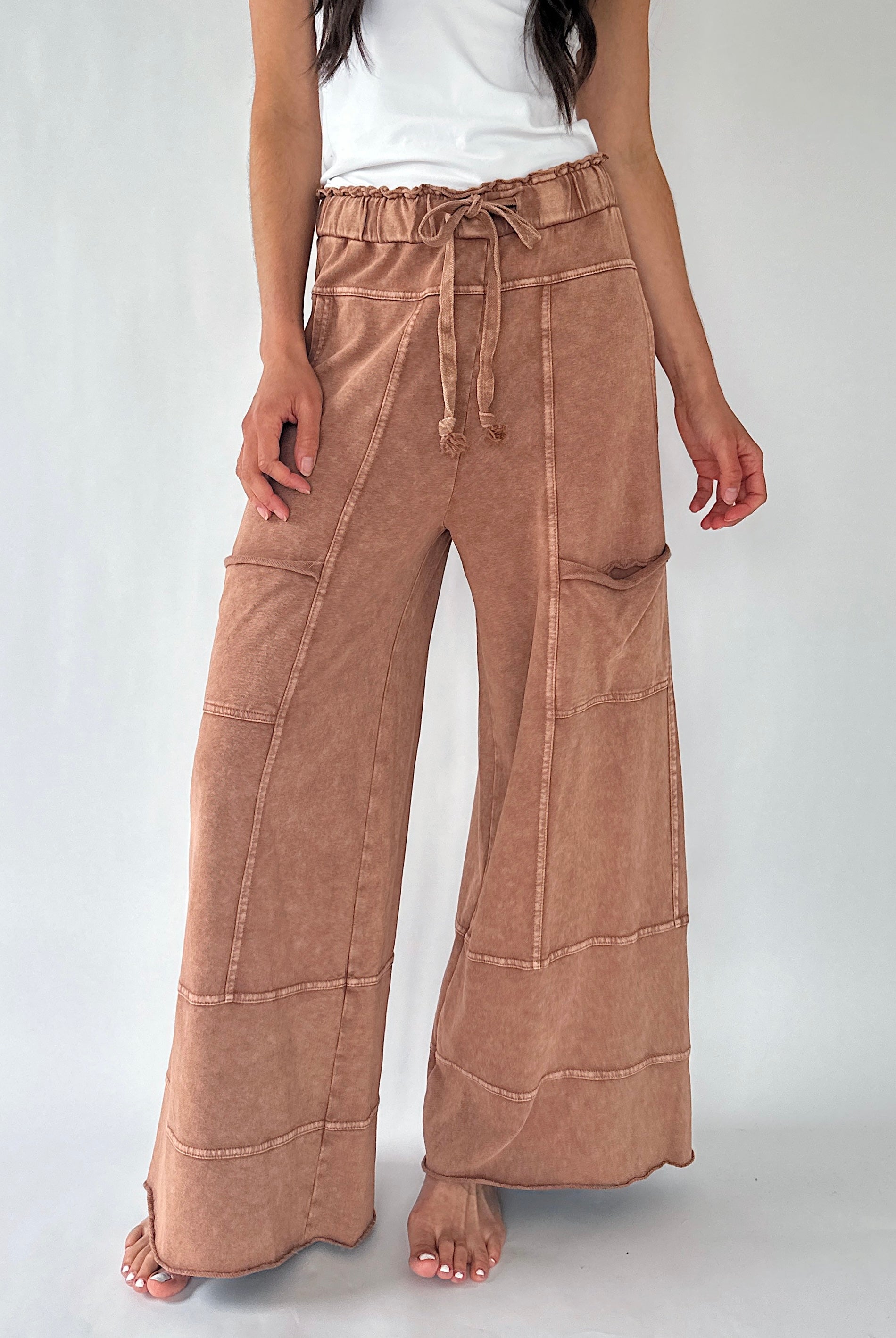 A model wearing a pair of wide-leg, high-waisted pants in a dusty tan or light brown color, styled with a white sleeveless top against a light gray studio background.