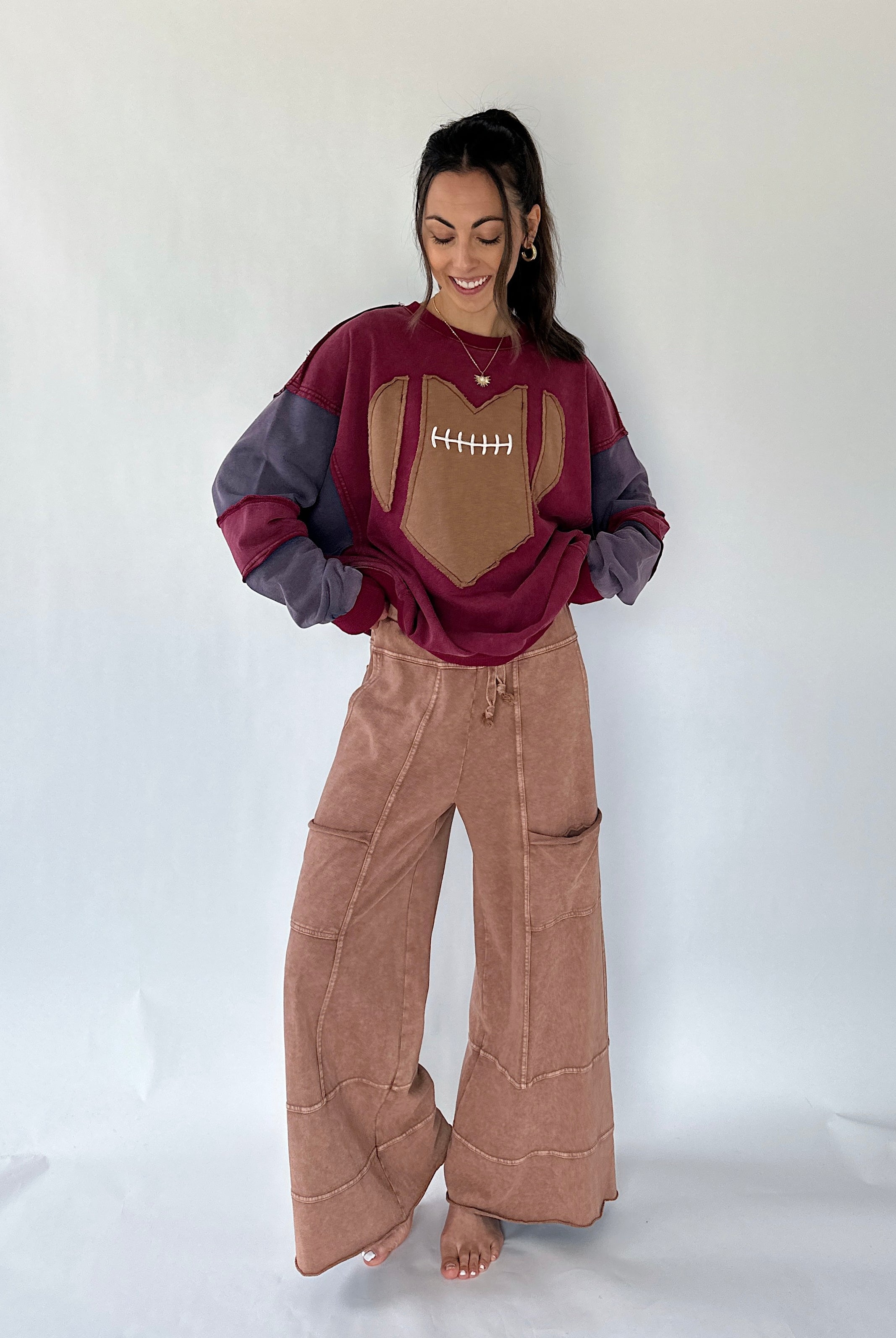 A woman modeling a football-themed sweatshirt and brown wide-leg pants against a plain white studio backdrop. The sweatshirt is burgundy and gray with a tan football patch on the chest, and the pants are a dusty rose-brown color.