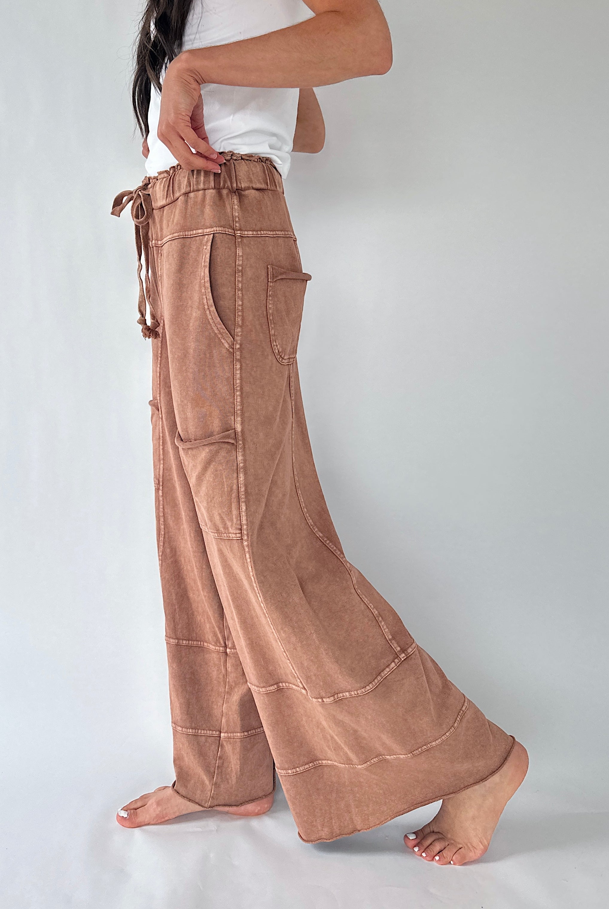 A person wearing loose, wide-leg brown pants with a drawstring waist and large patch pockets, standing barefoot against a plain light gray studio background.