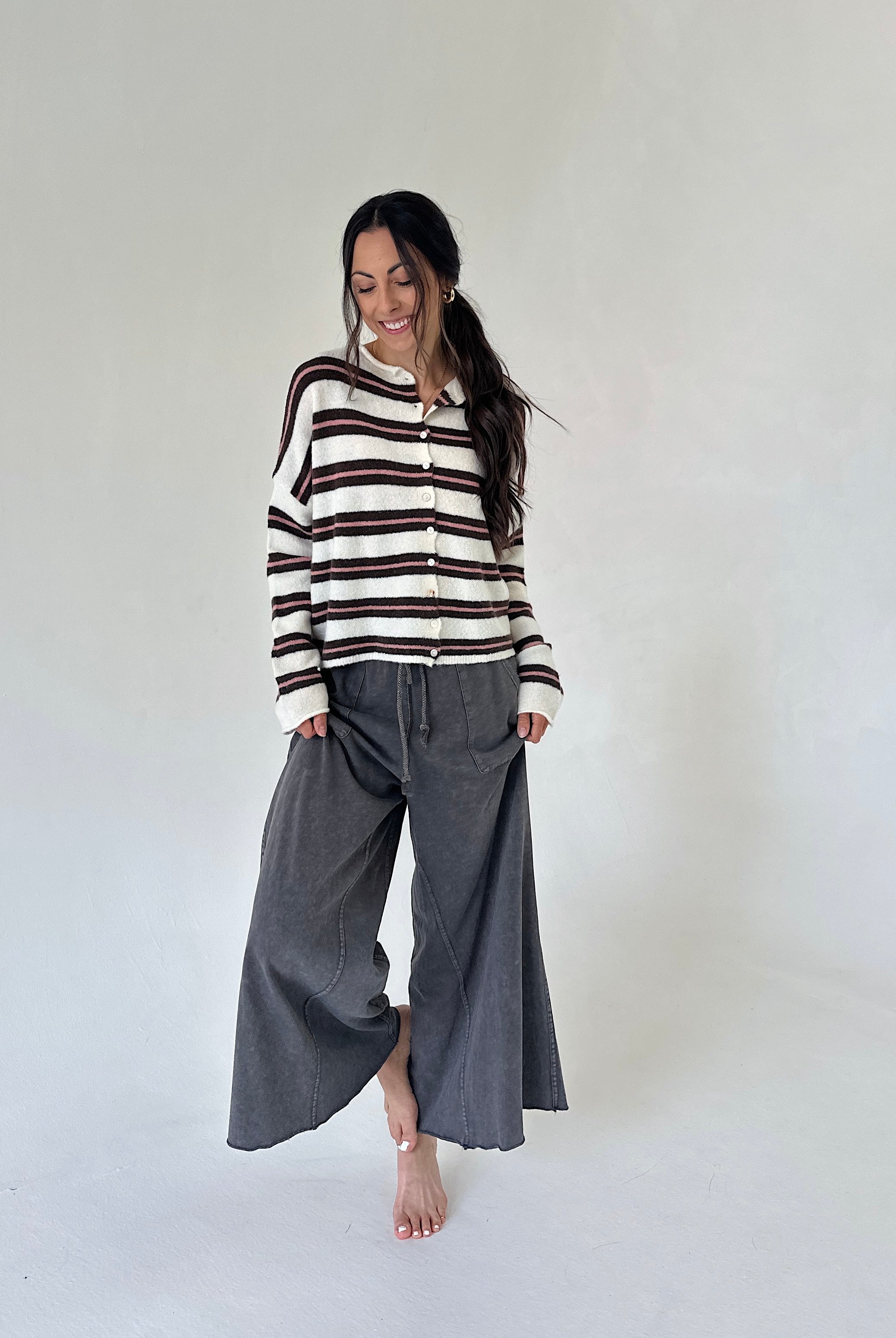 A woman stands barefoot against a plain light gray studio background, wearing a cream knit cardigan with dark brown horizontal stripes and loose dark gray wide-leg pants.