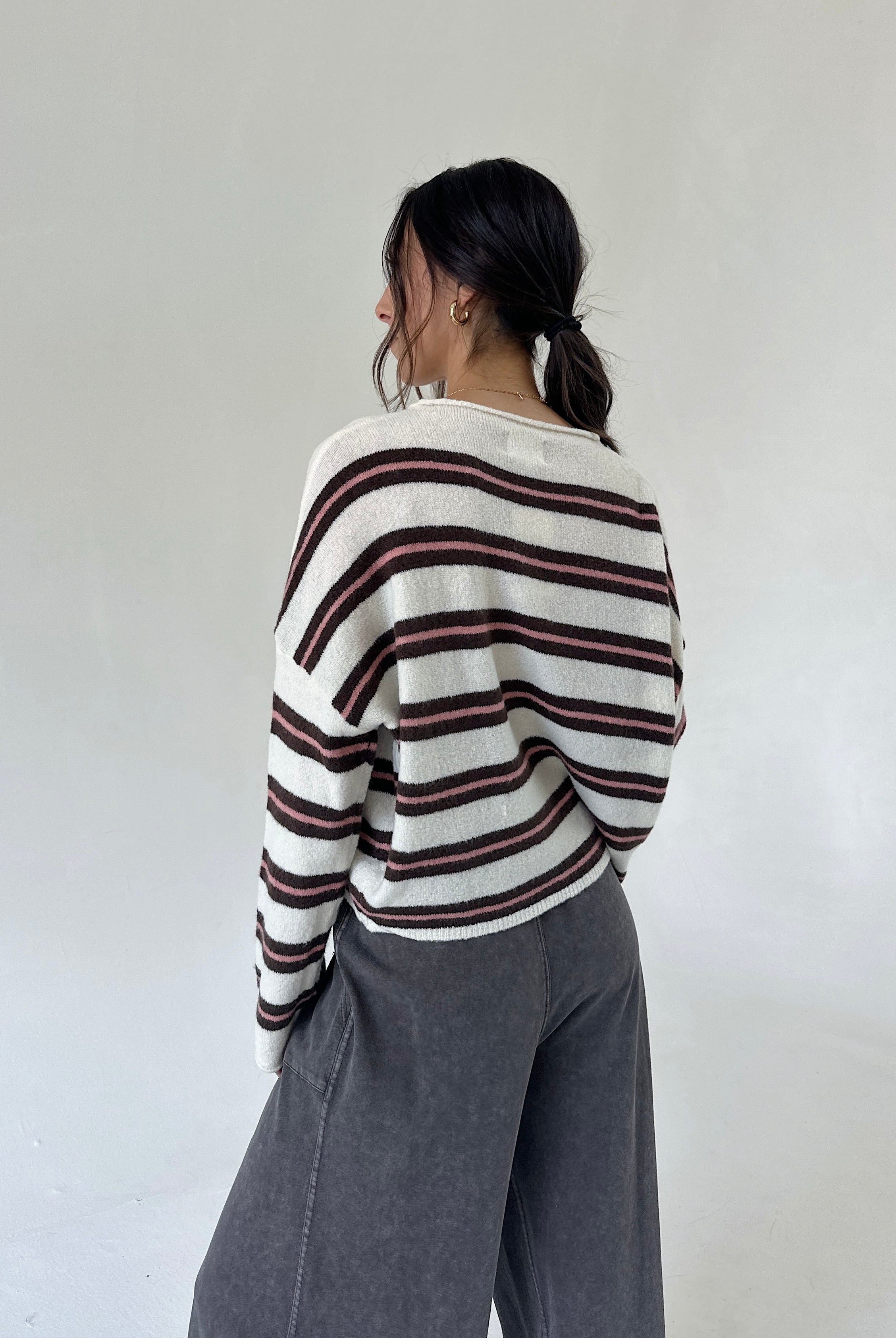A woman is shown from behind wearing a cream-colored knit sweater with dark brown and pink horizontal stripes, paired with wide-leg gray pants, against a plain off-white studio background.