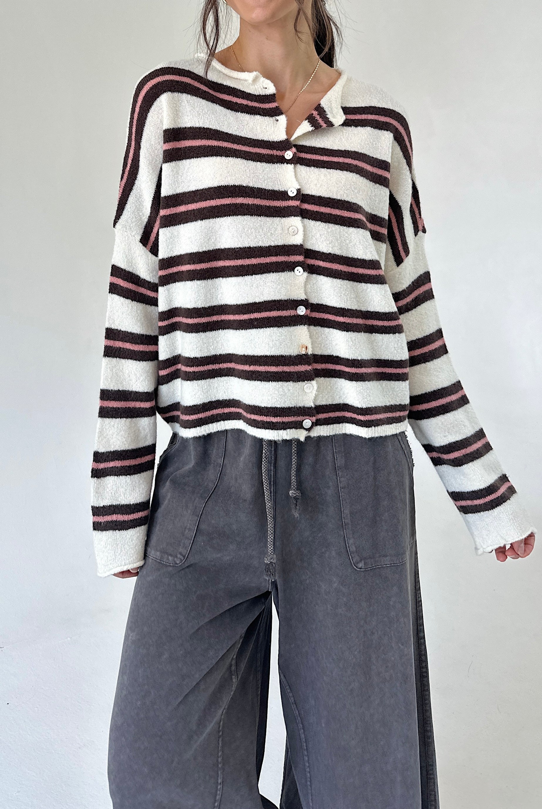 A woman wearing a cream knit button-up cardigan with dark brown and pink horizontal stripes, paired with wide-leg charcoal gray pants, against a plain light gray background.