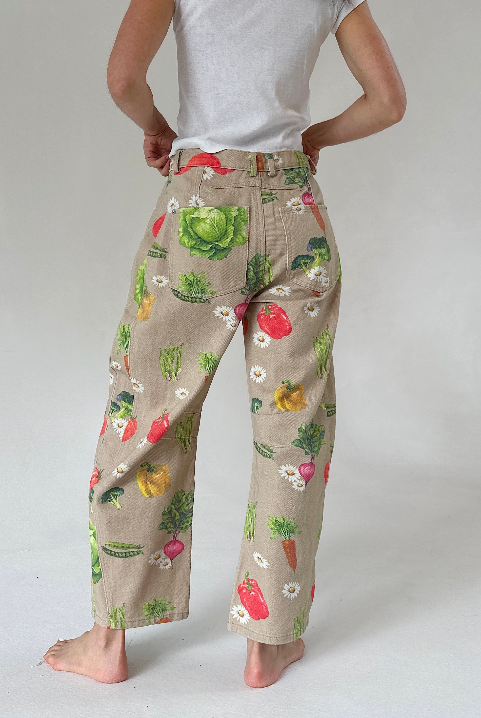 A person shown from the back wearing beige wide-leg pants with a colorful vegetable print, paired with a white t-shirt, against a light gray studio background.