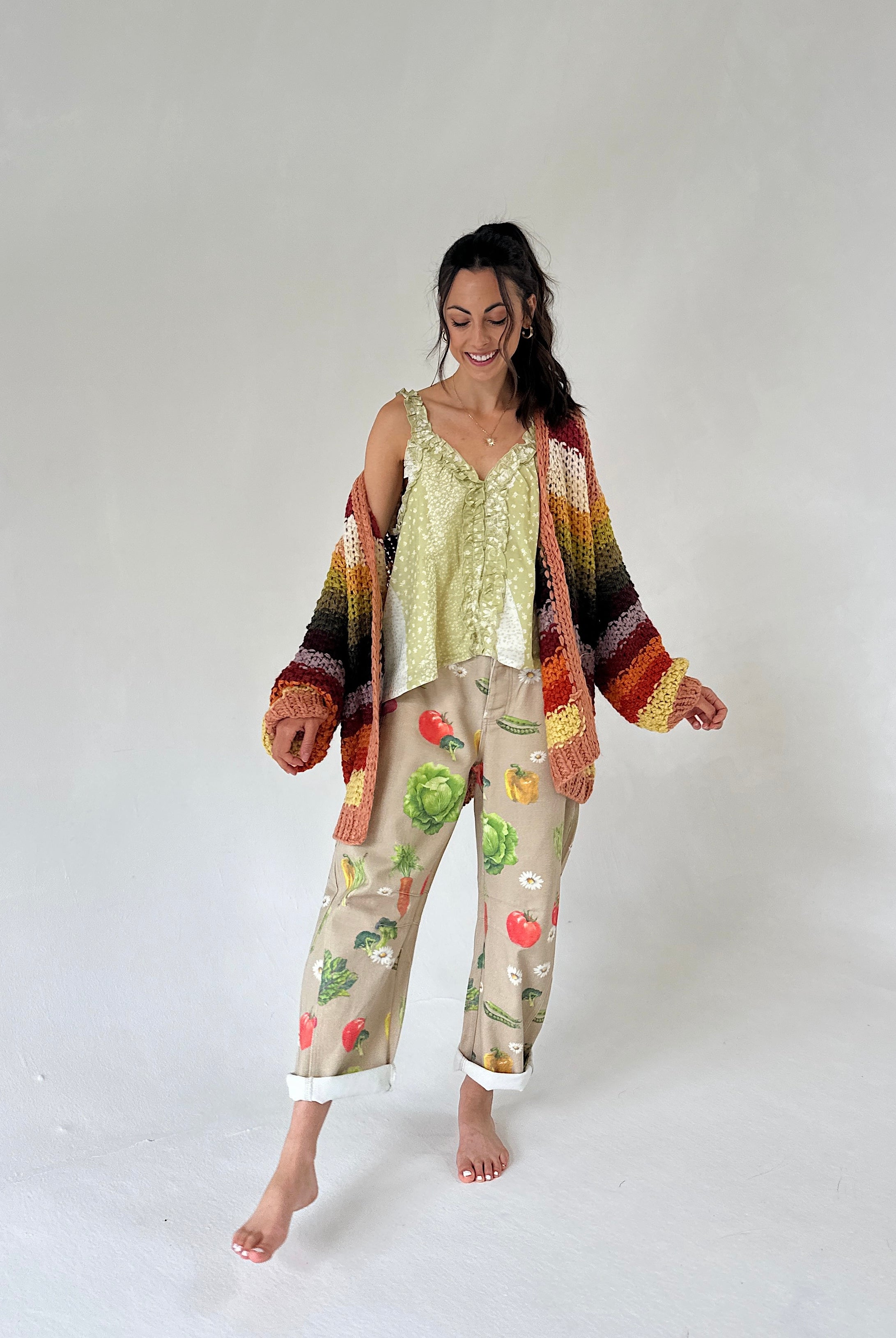 A woman modeling a light green printed tank top and beige cropped pants with a colorful vegetable print, styled with a multicolor striped knit cardigan, against a plain light gray studio background.