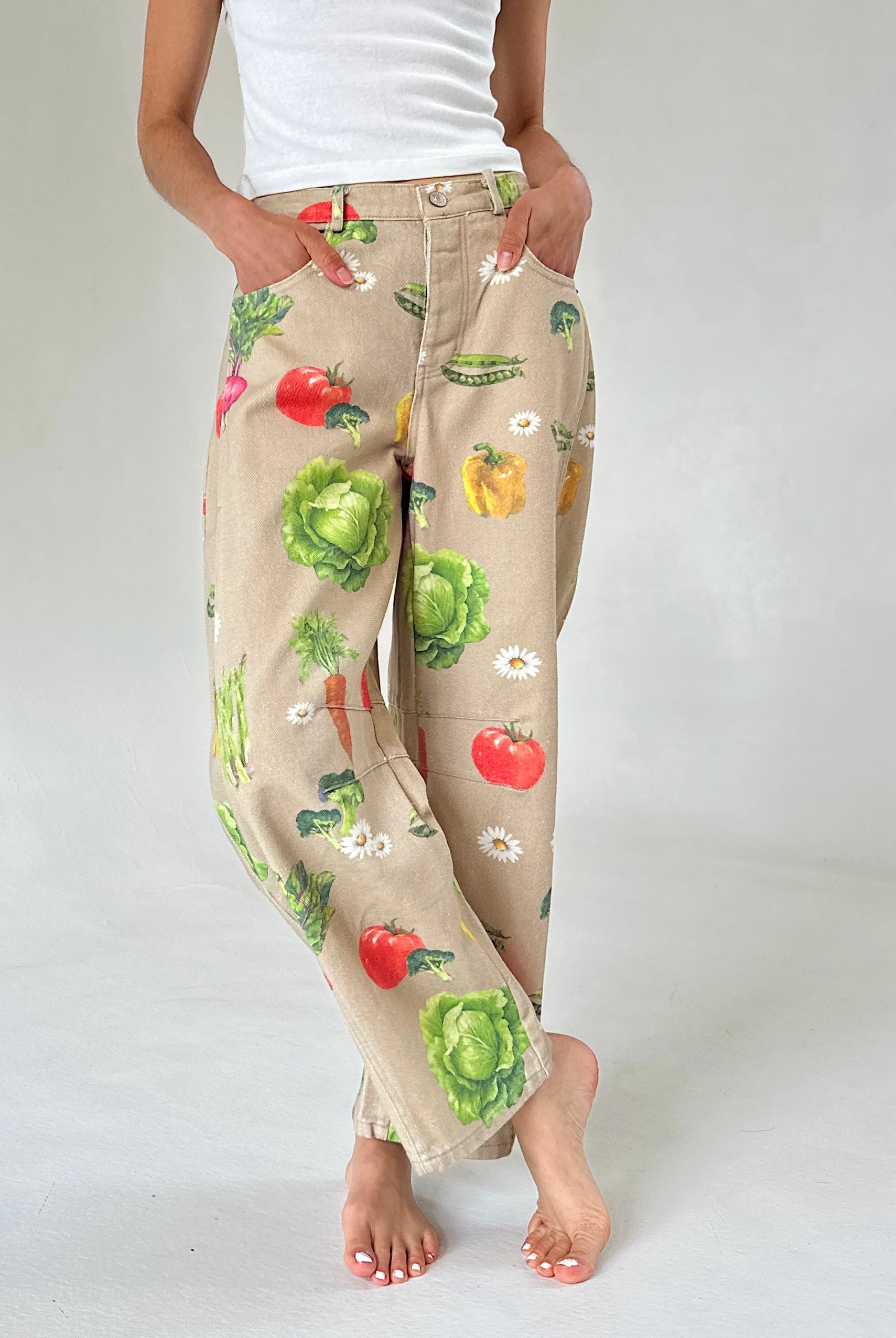 A person wearing beige wide-leg pants with a colorful vegetable print, legs crossed, paired with a white tank top, shown against a light gray studio background.