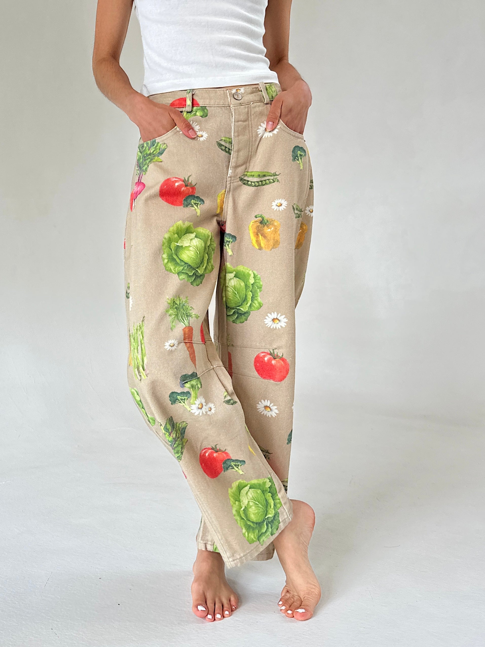 A person wearing beige wide-leg pants with a colorful vegetable print, legs crossed, paired with a white tank top, shown against a light gray studio background.