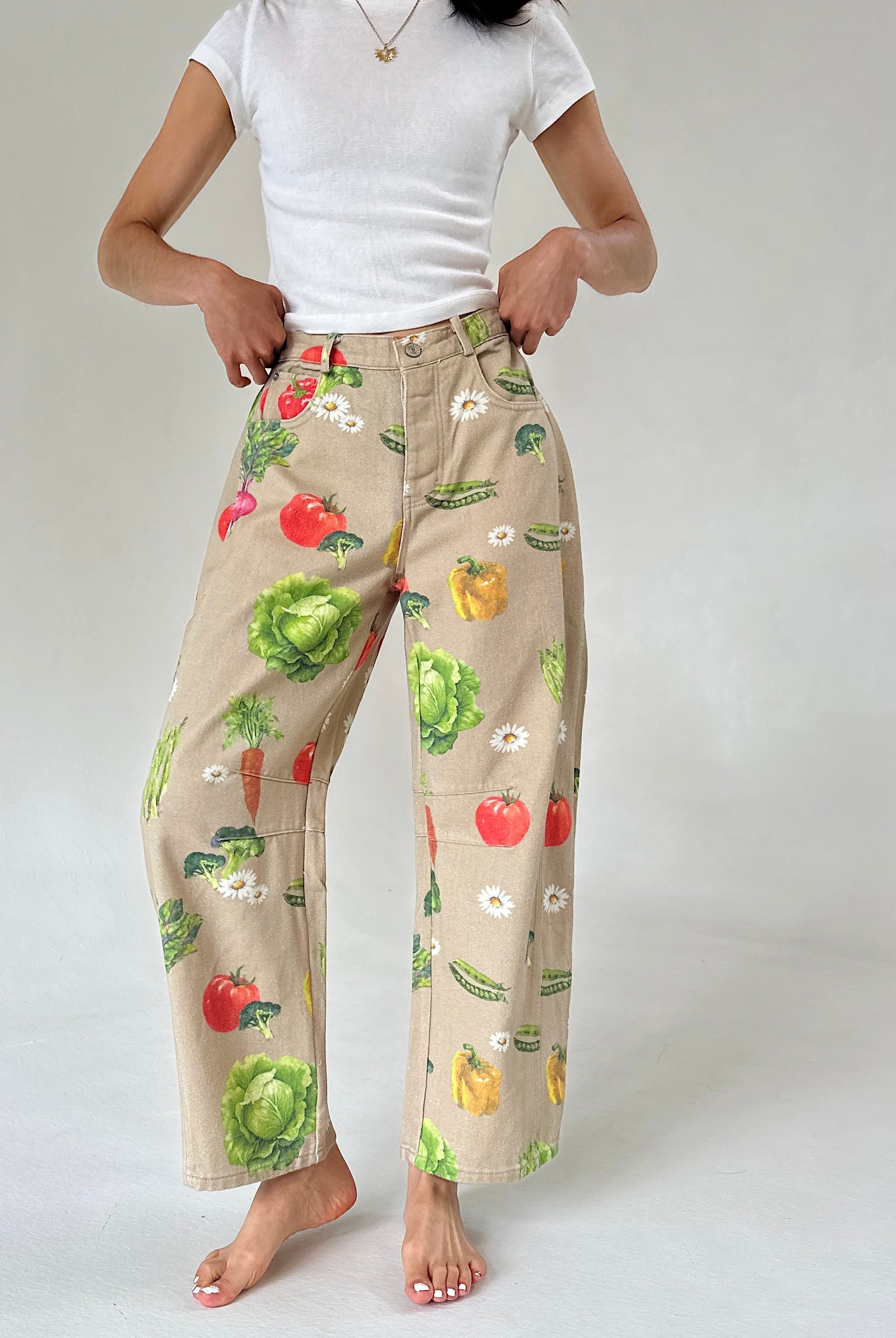 A person wearing beige wide-leg barrel pants with a colorful vegetable print, paired with a white t-shirt, standing against a light gray studio background.