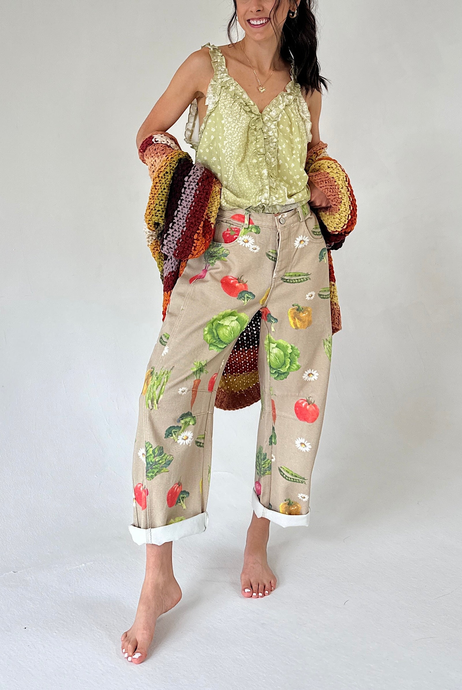 A woman modeling loose beige cropped pants with a colorful vegetable print, paired with a light green printed tank and holding a chunky knit cardigan.