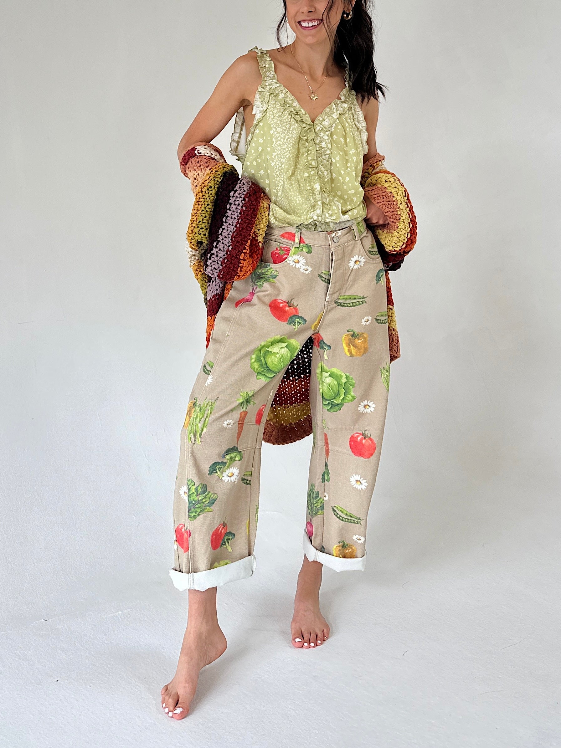 A woman modeling loose beige cropped pants with a colorful vegetable print, paired with a light green printed tank and holding a chunky knit cardigan.