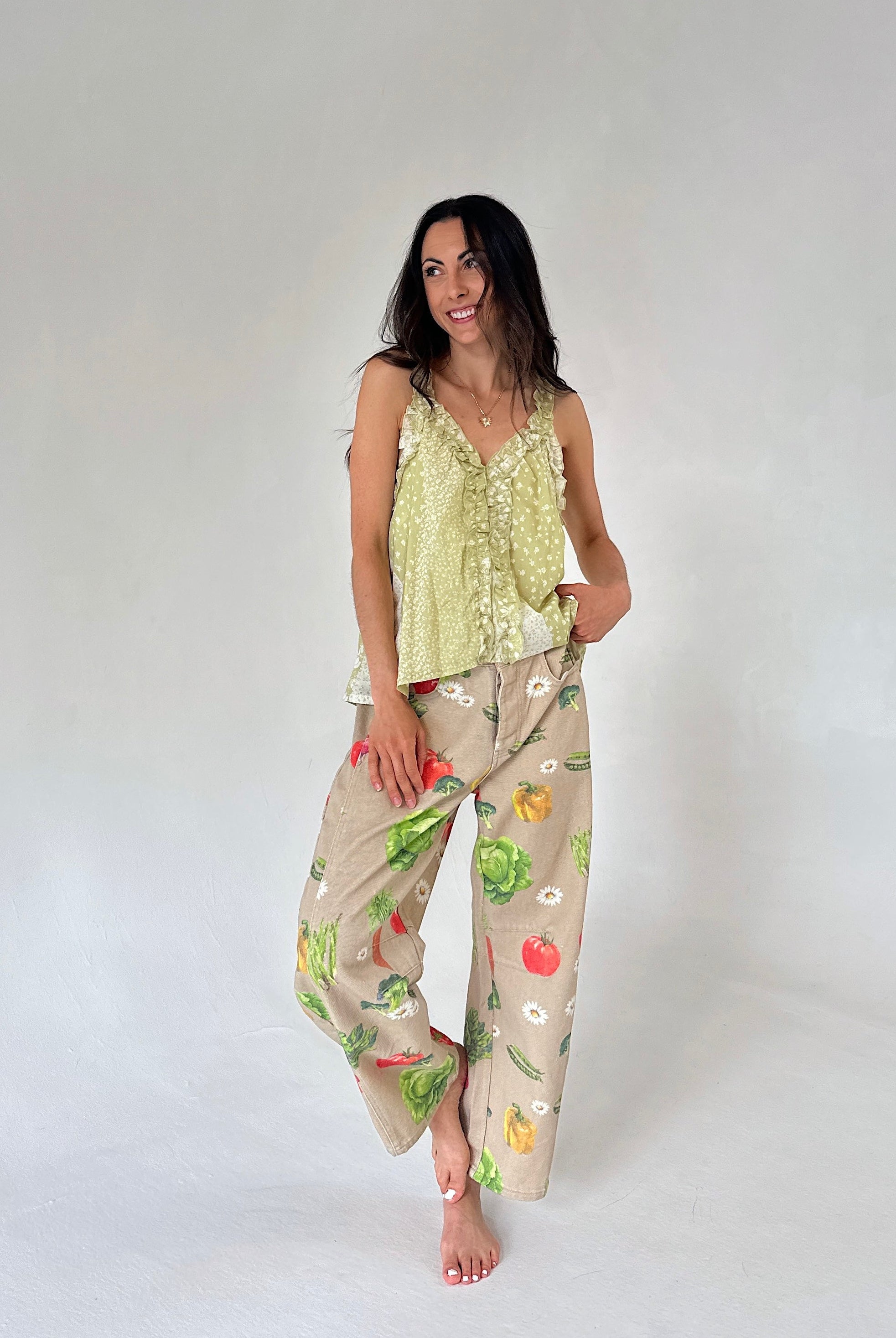 A woman modeling a light yellow-green ruffled camisole top and barrel pants with a colorful vegetable print, set against a plain light gray studio background.