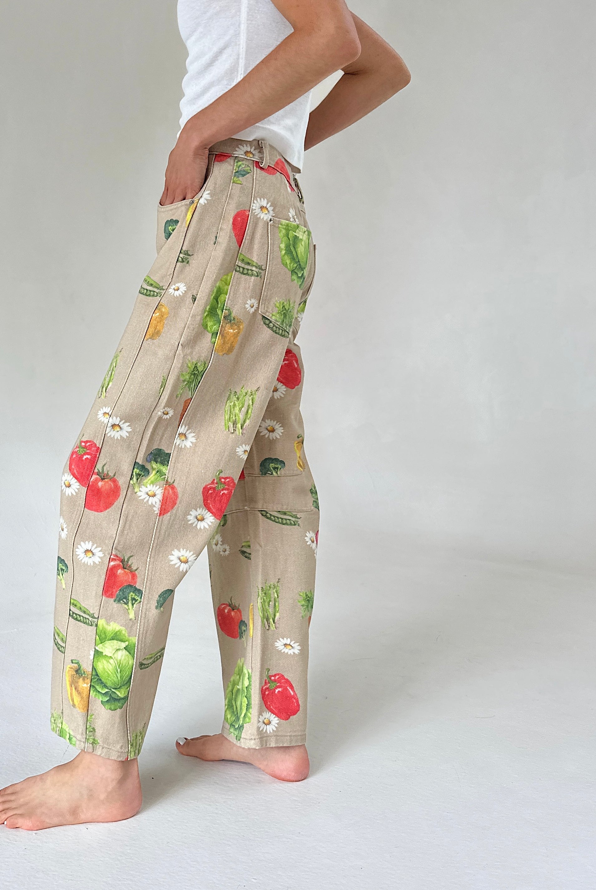 A person wearing beige wide-leg pants with a colorful vegetable print, standing barefoot against a light gray studio background.