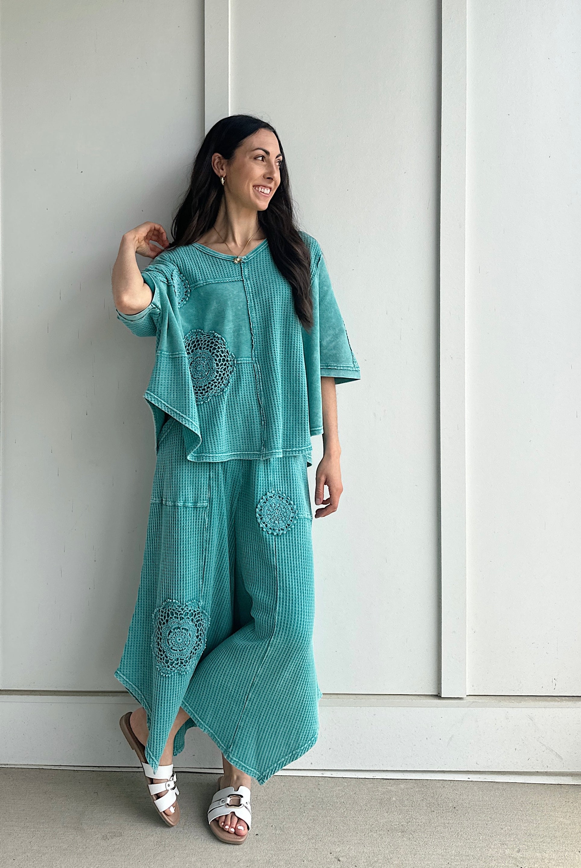Woman wearing a teal outfit with patterns against a white paneled wall.
