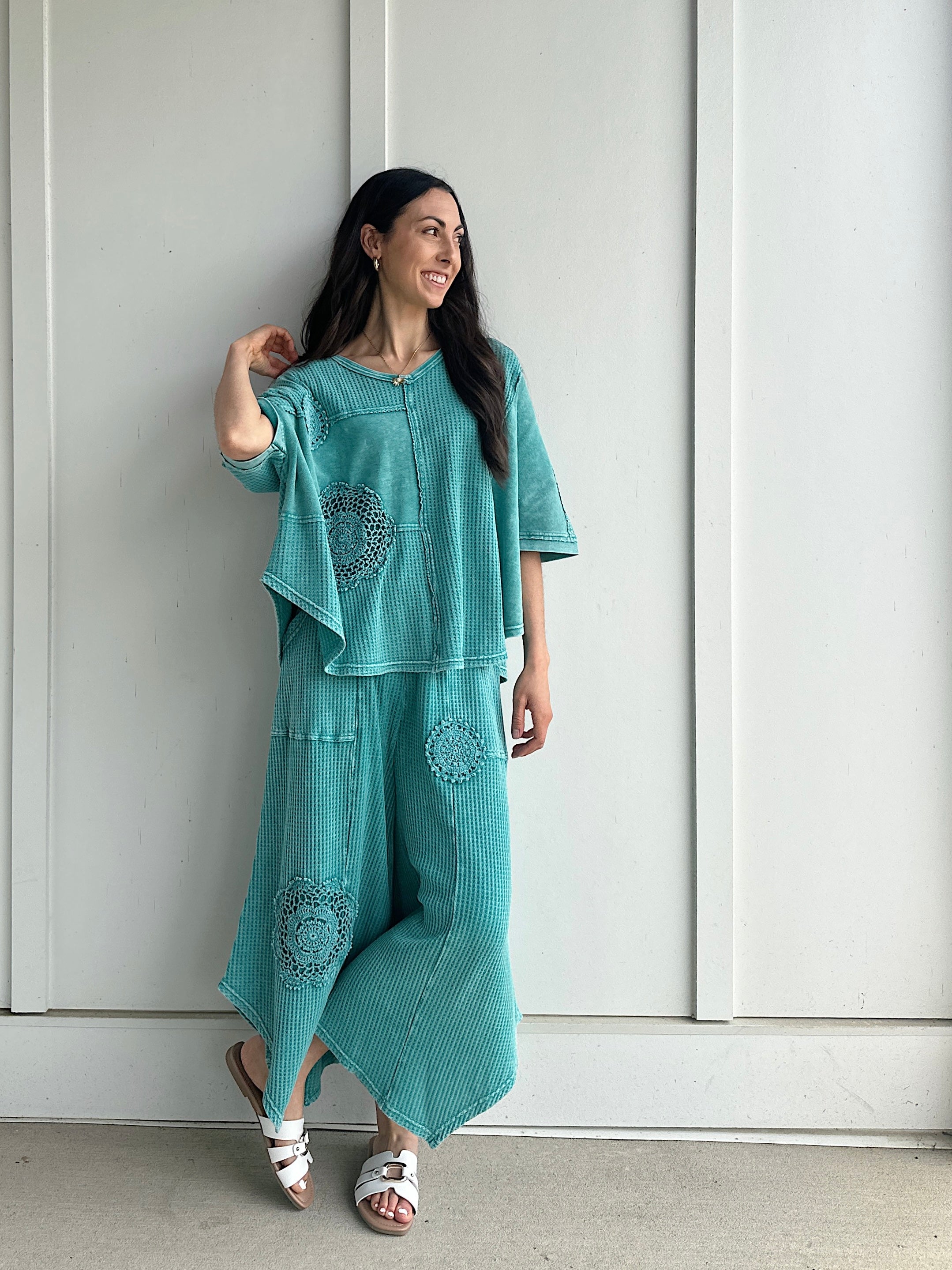 Woman wearing a teal outfit with patterns against a white paneled wall.