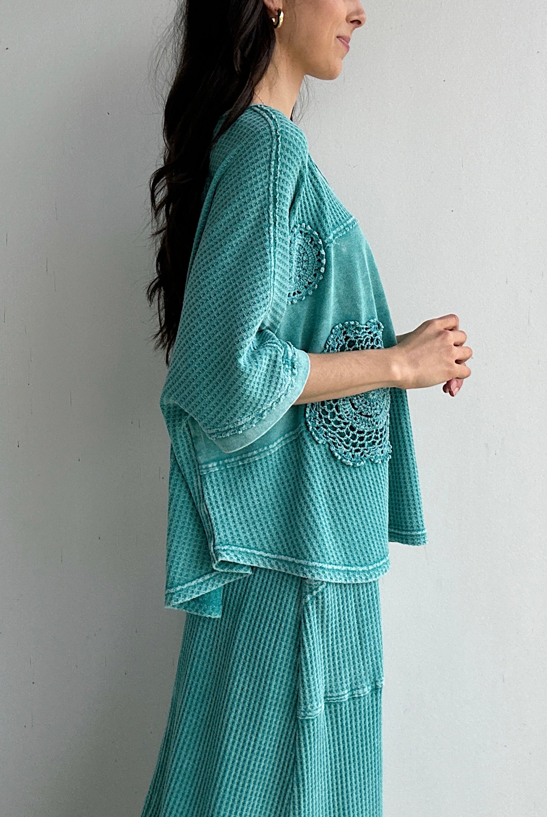 Woman wearing a teal textured outfit with lace details against a plain background showing side