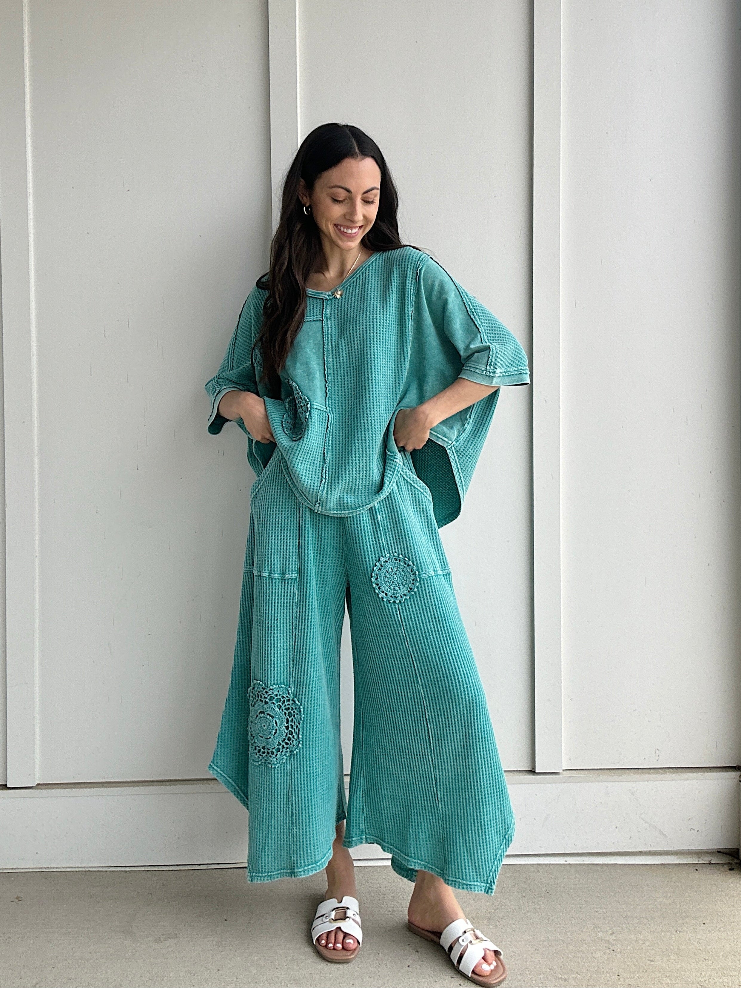 Woman wearing a teal outfit with a matching top and wide-leg pants against a white wall.