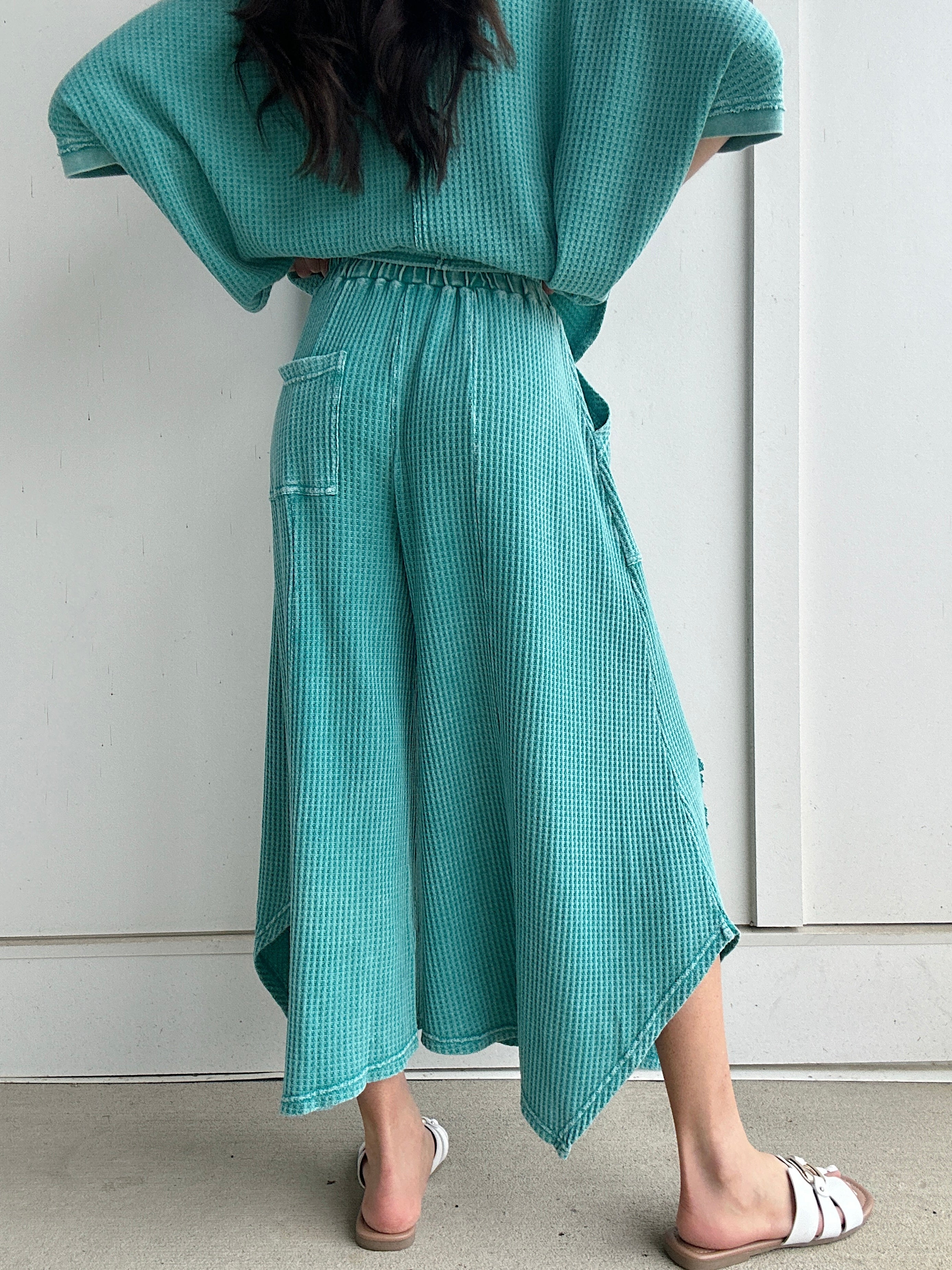 Person wearing a teal outfit with a matching wide leg pants against a white wall showing the back of the pants