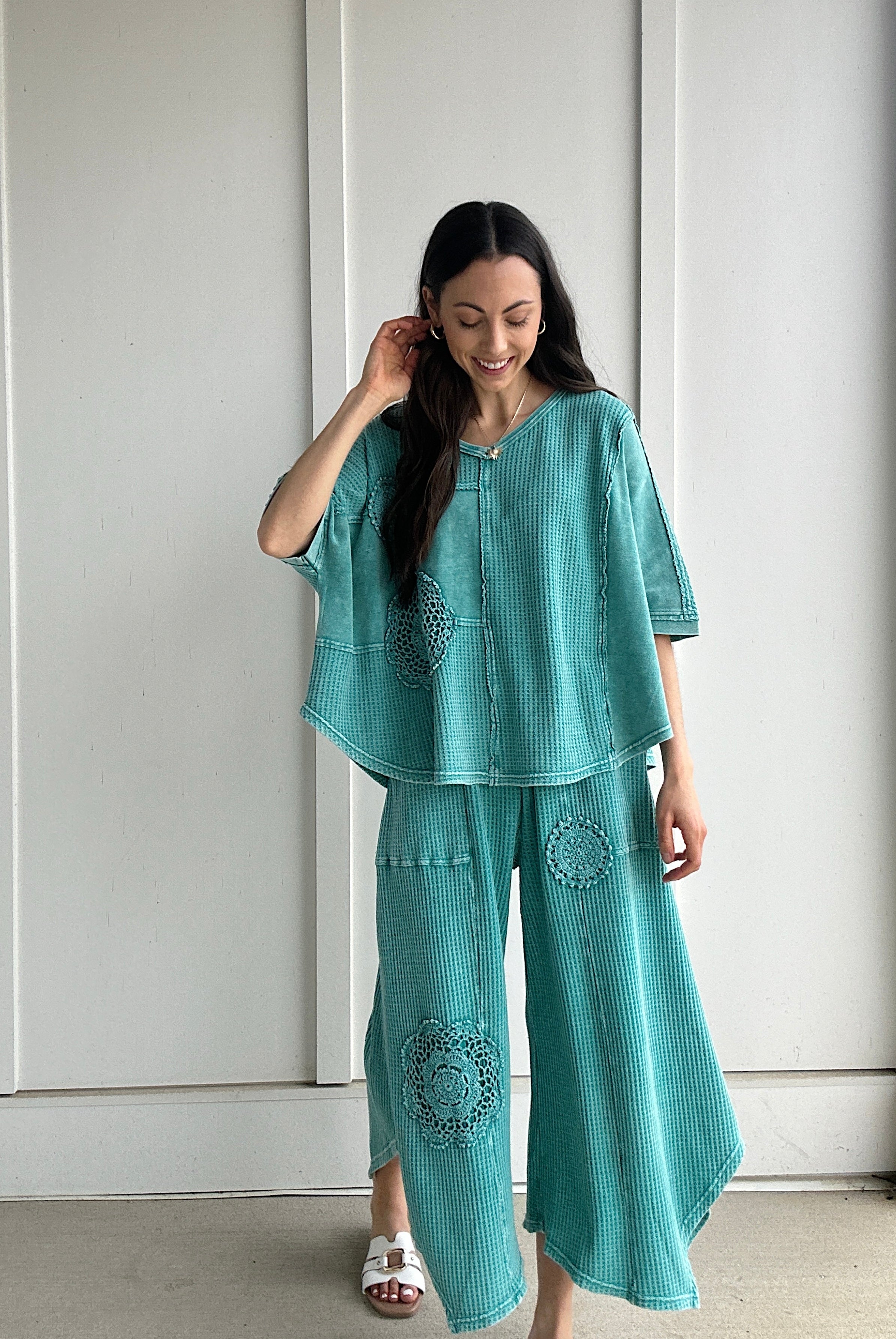 Woman wearing a teal outfit with crochet patterns against a white paneled wall.