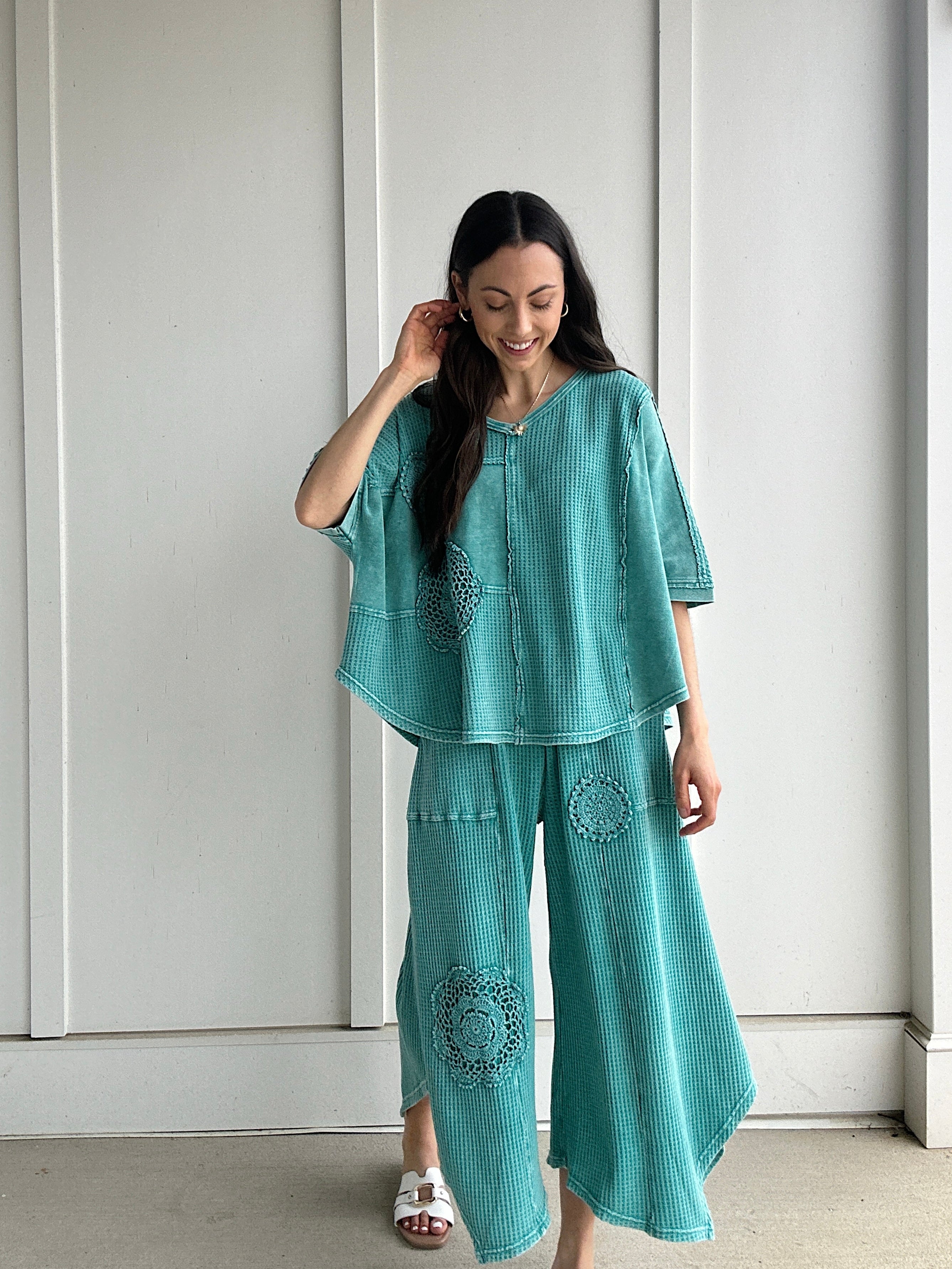 Woman wearing a teal outfit with crochet patterns against a white paneled wall.
