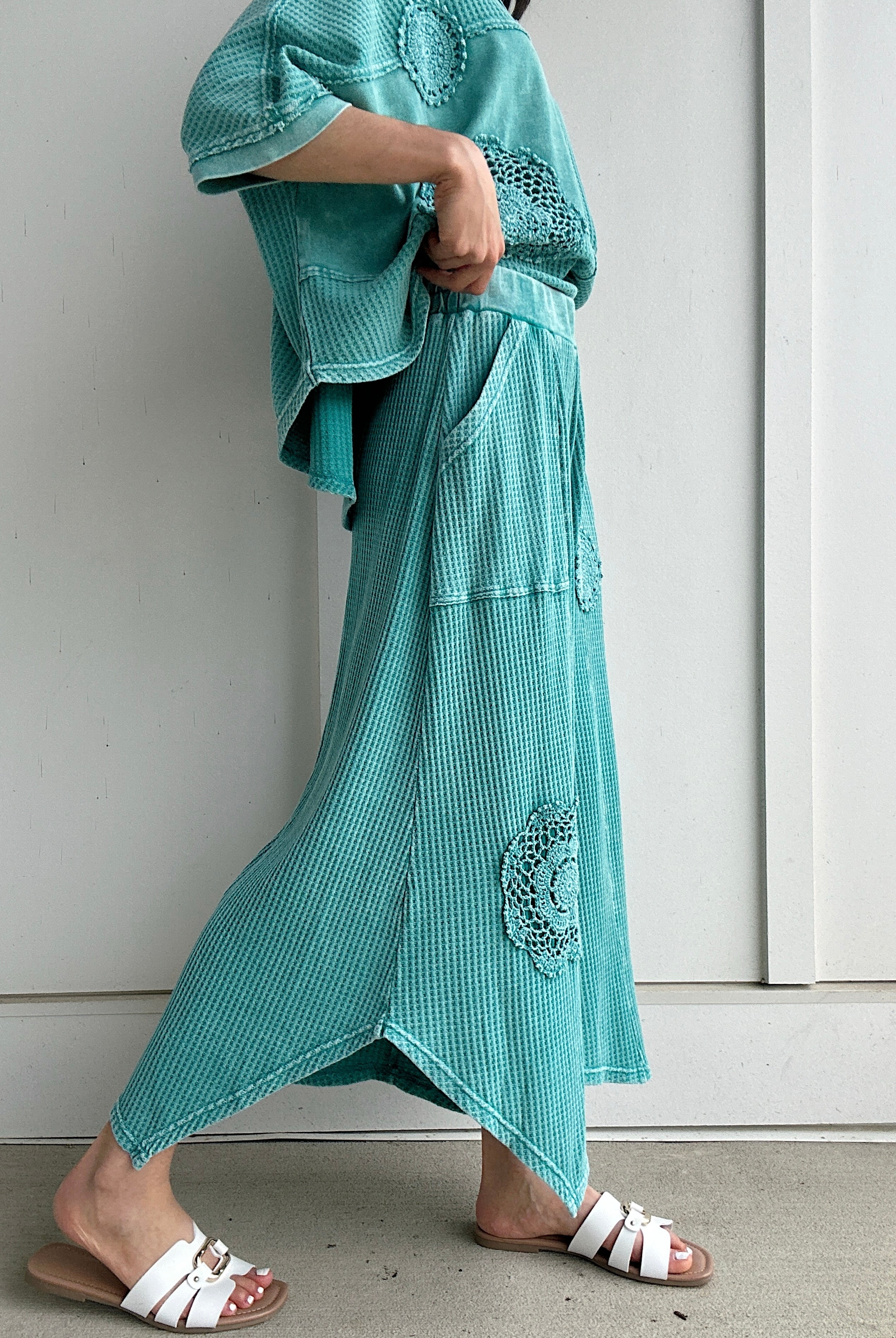 Person wearing a teal outfit with embroidered details against a white wall showing side