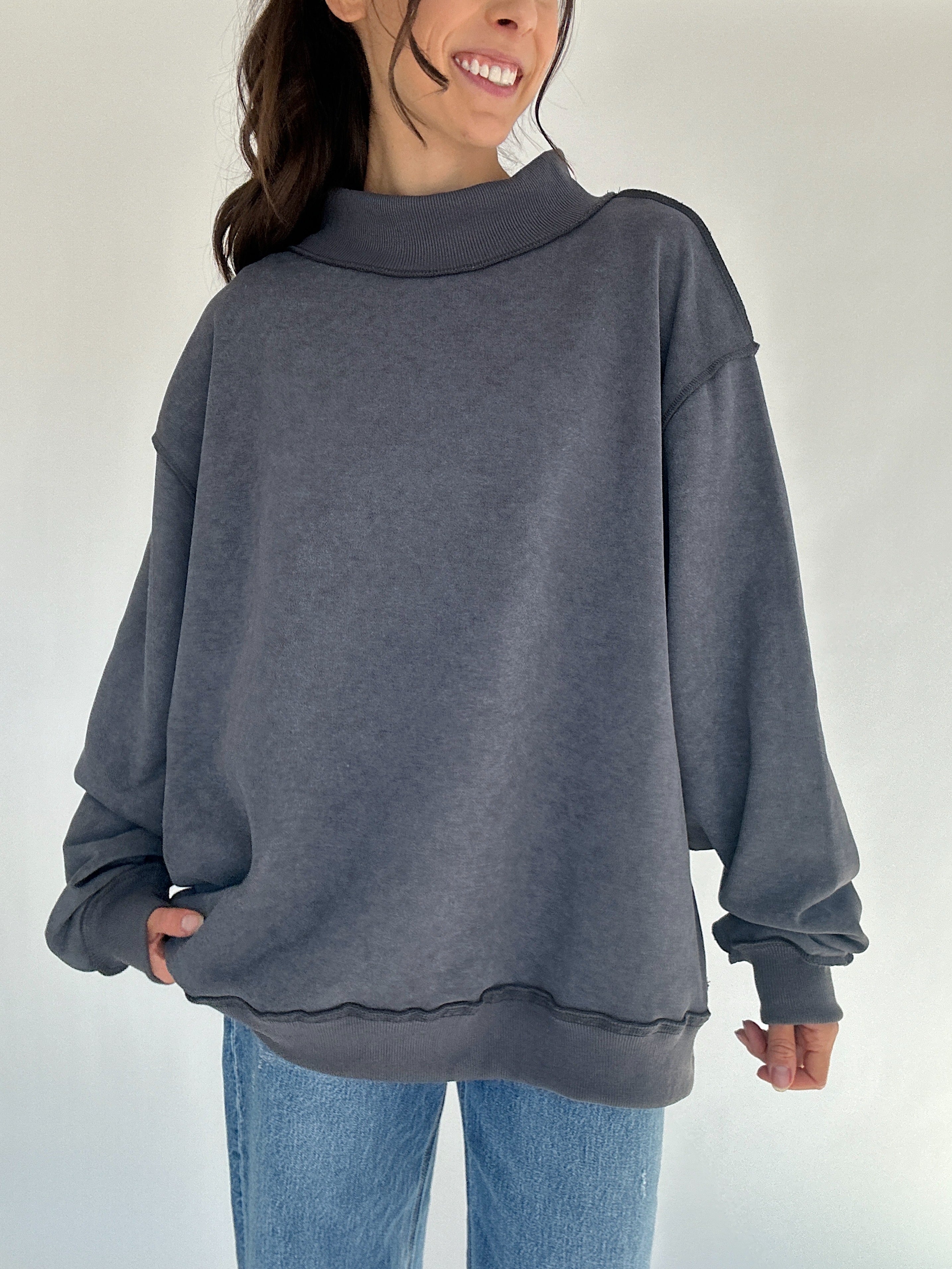 Person wearing a gray oversized mock neck sweatshirt and blue jeans on a plain background