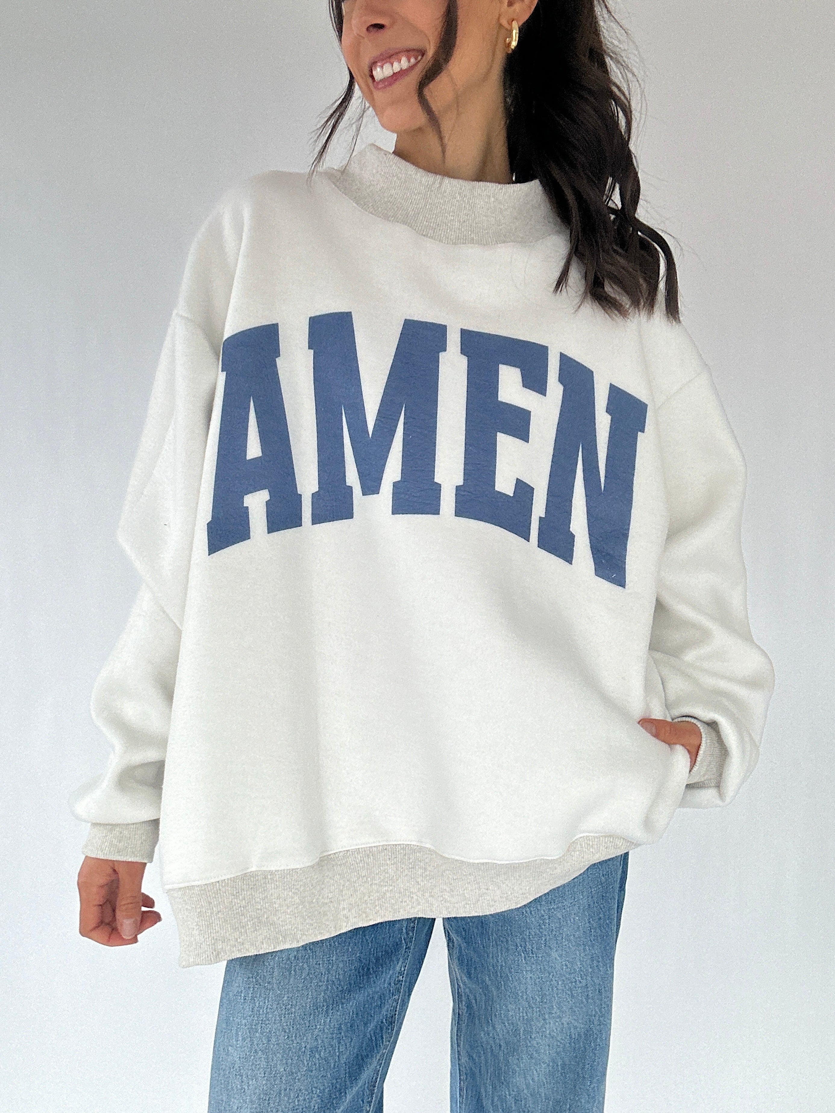Person wearing a white sweatshirt with 'AMEN' printed in blue on a plain background