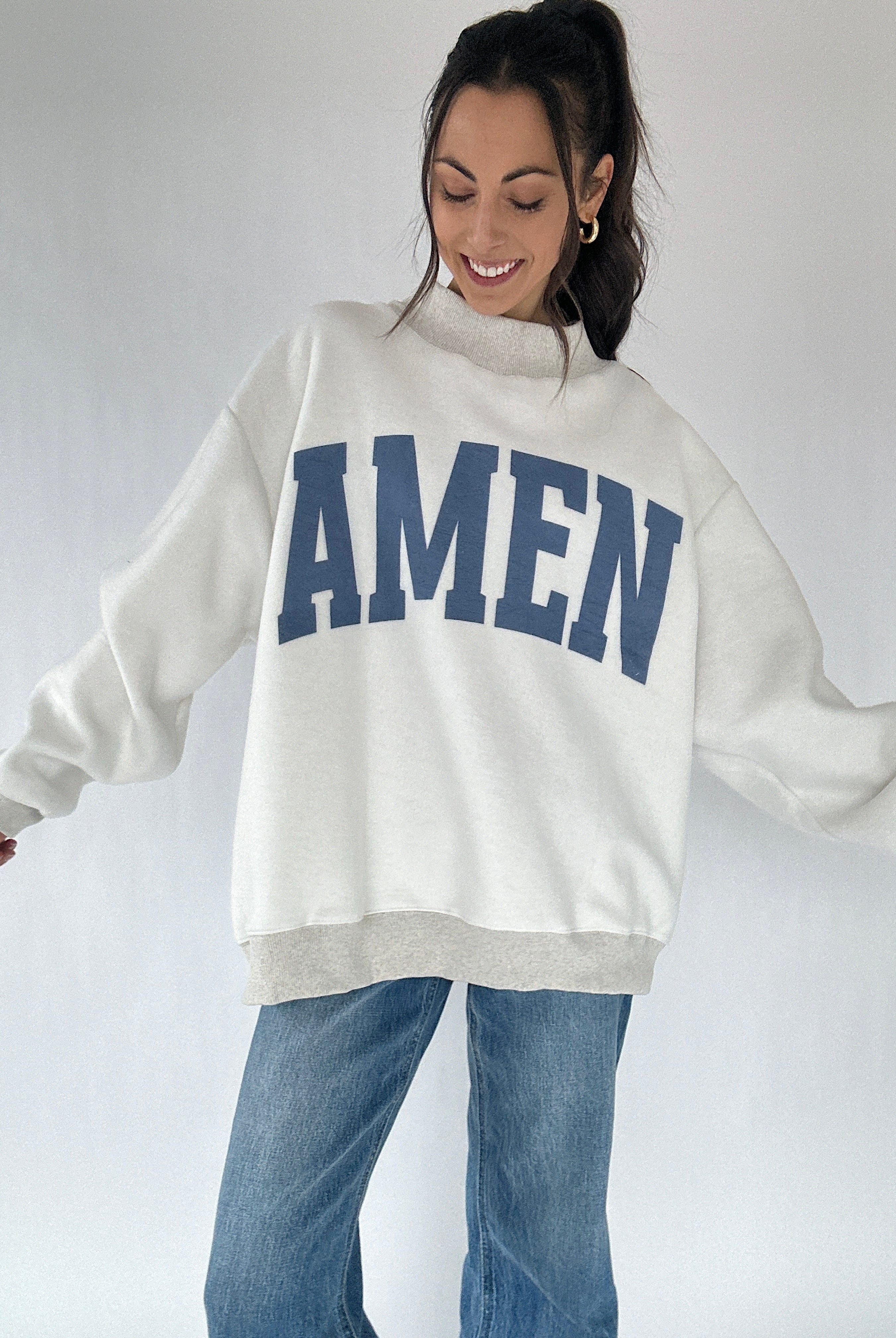 Woman wearing a white sweatshirt with 'AMEN' printed in blue on a plain background