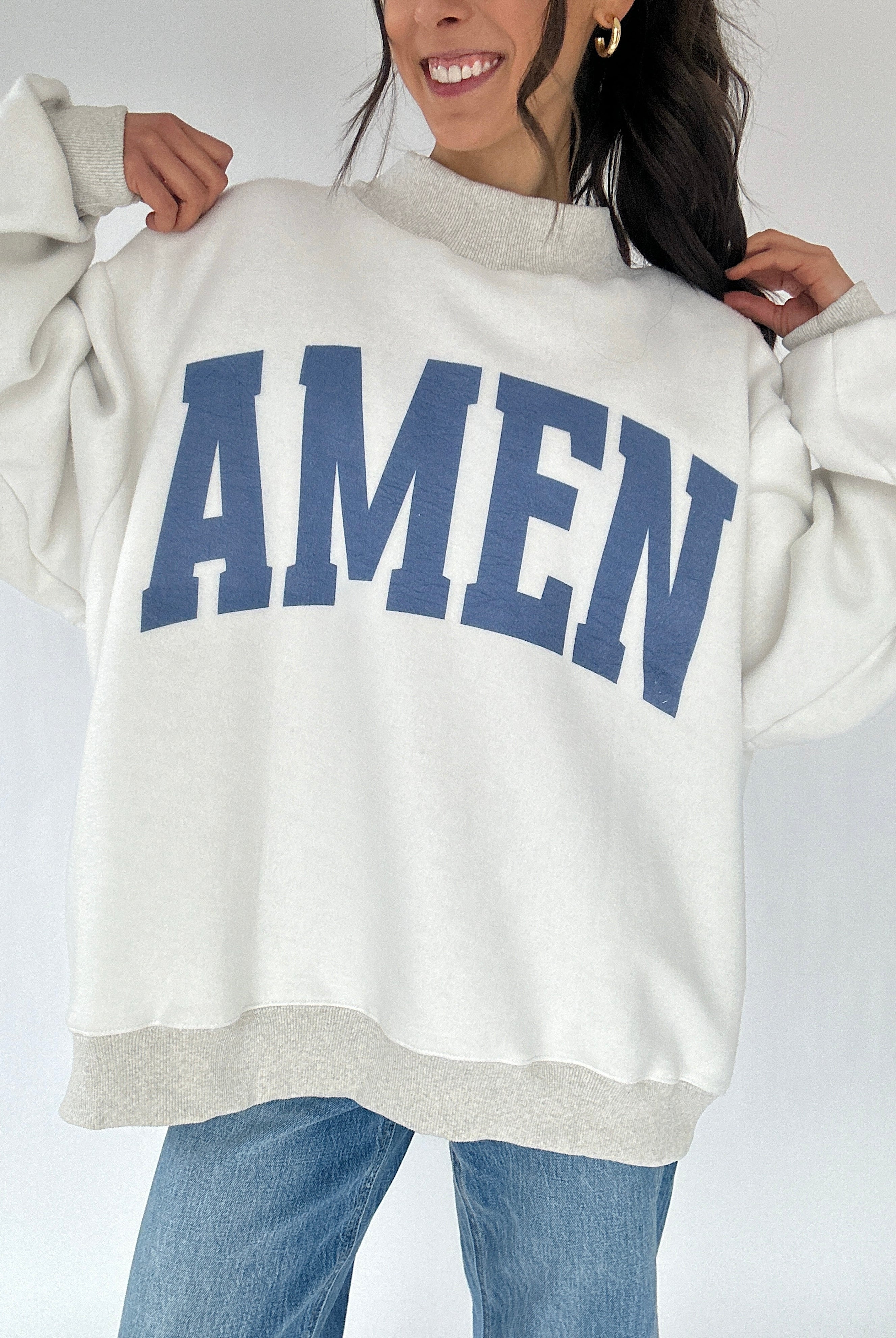 Person wearing a white sweatshirt with 'AMEN' printed in blue on a plain background