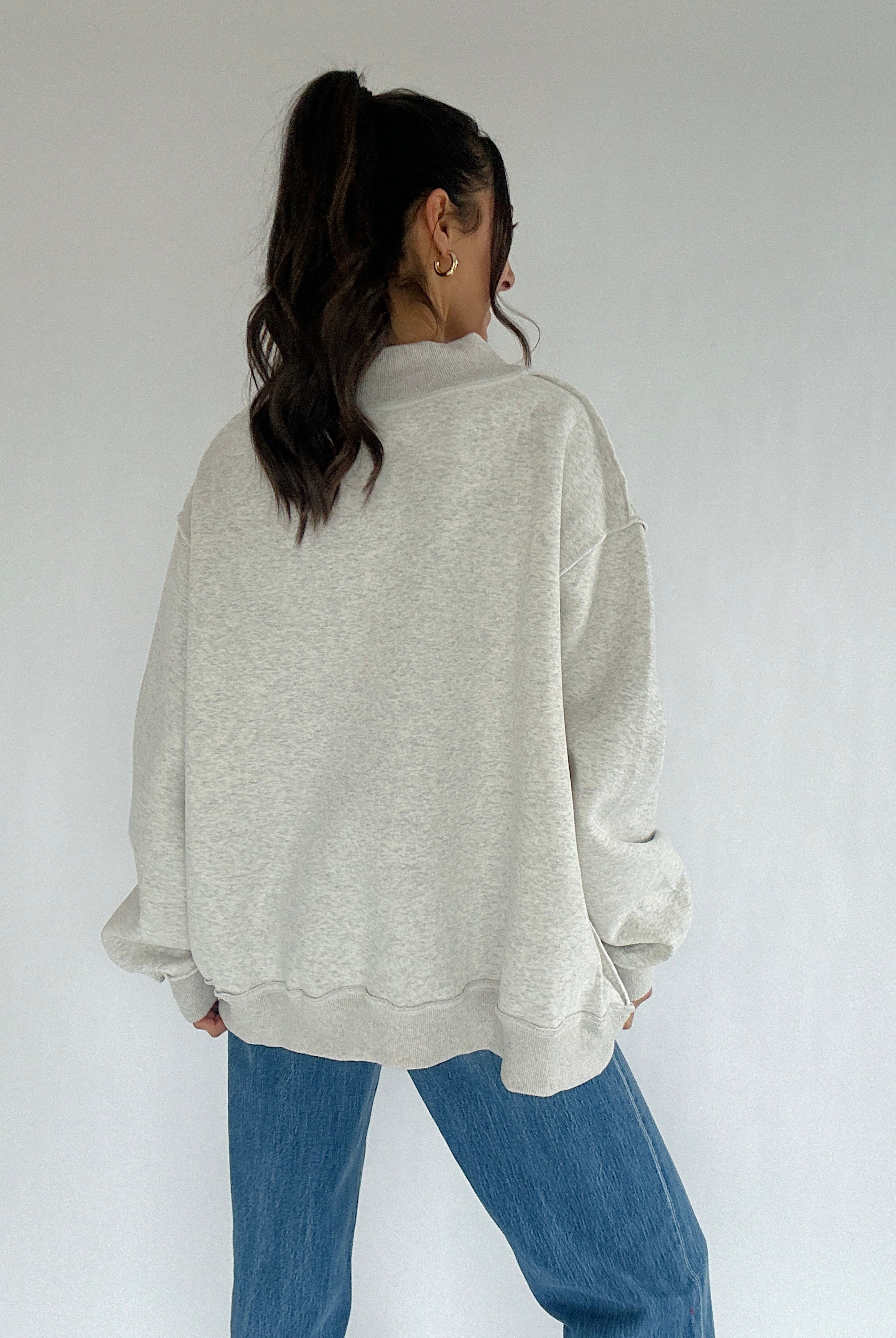 Person wearing a light gray sweatshirt and blue jeans against a white background showing back