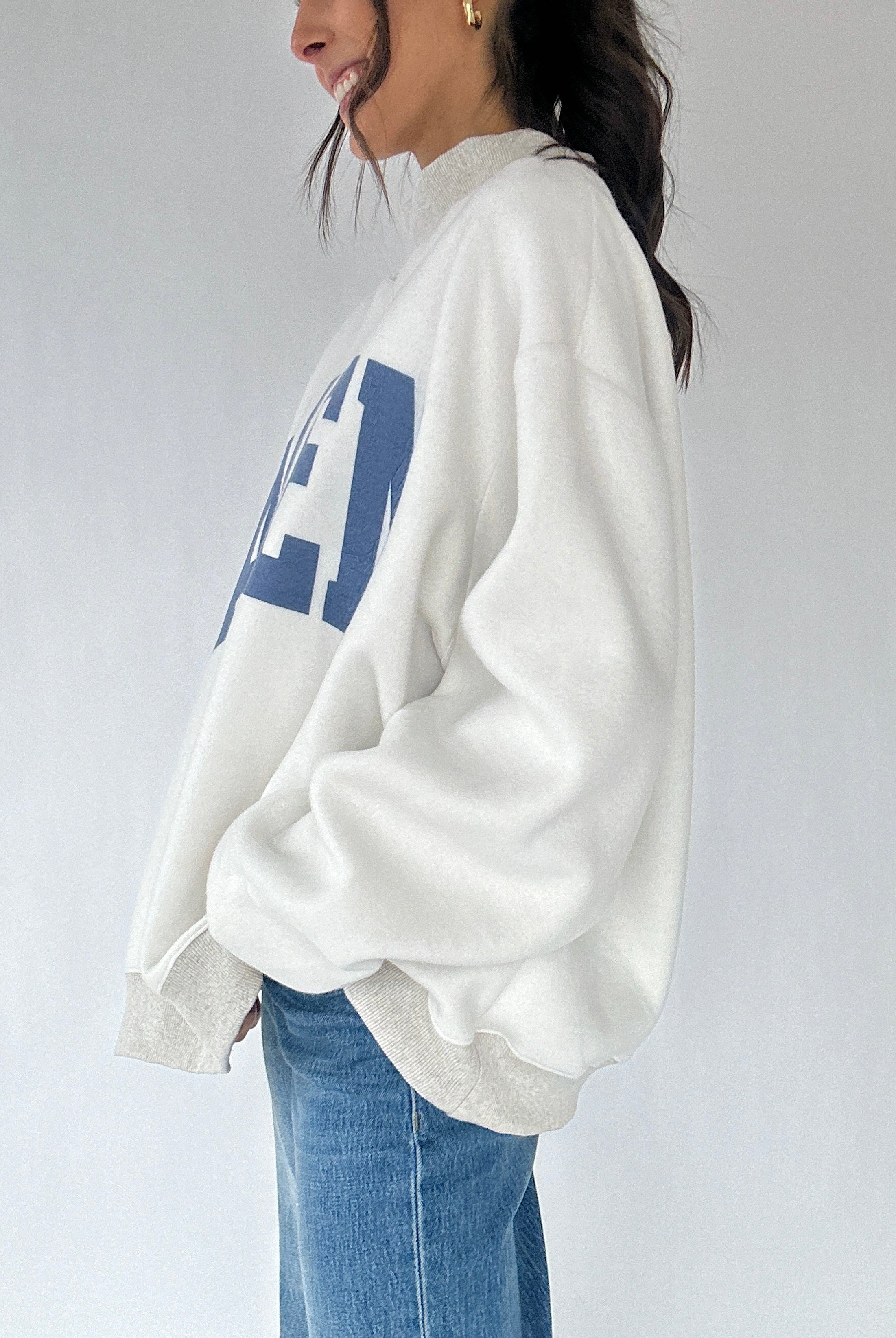 Person wearing a white sweatshirt with blue text and blue jeans on a plain background showing the side