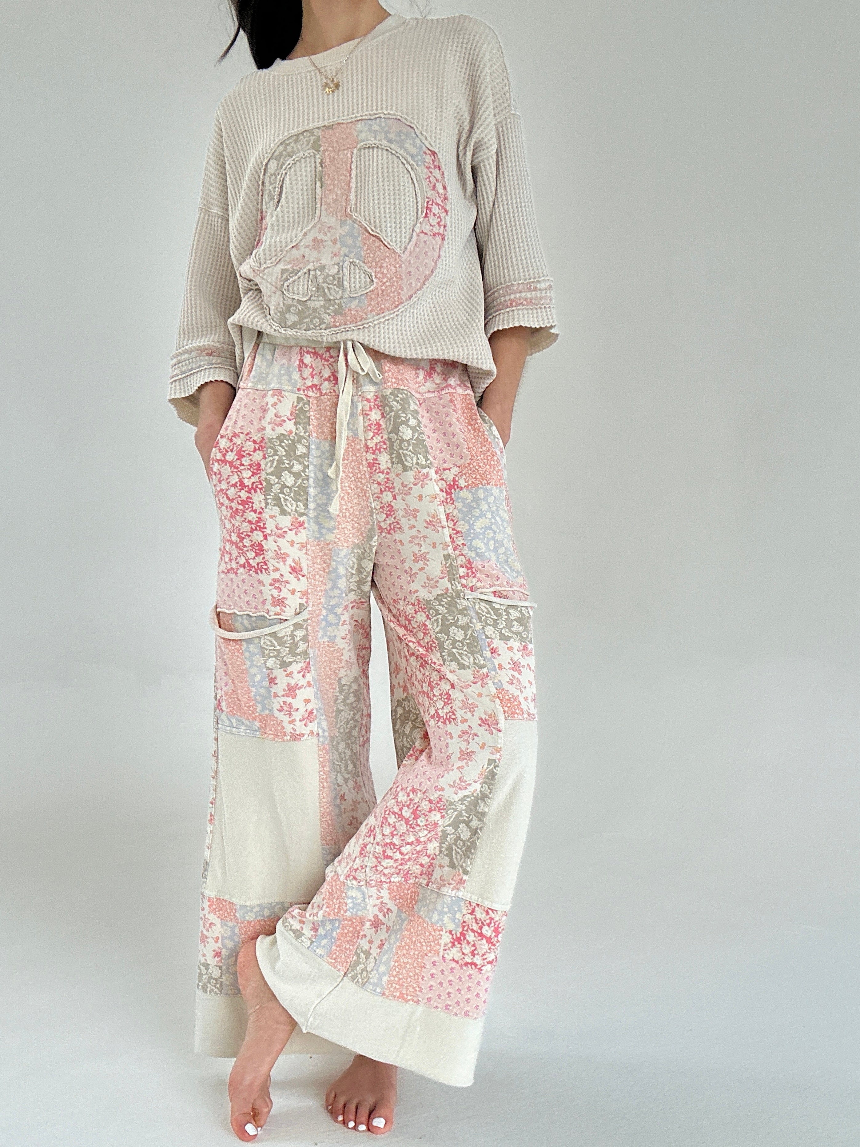 Person wearing a beige sweater with a peace symbol and floral pants on a plain background