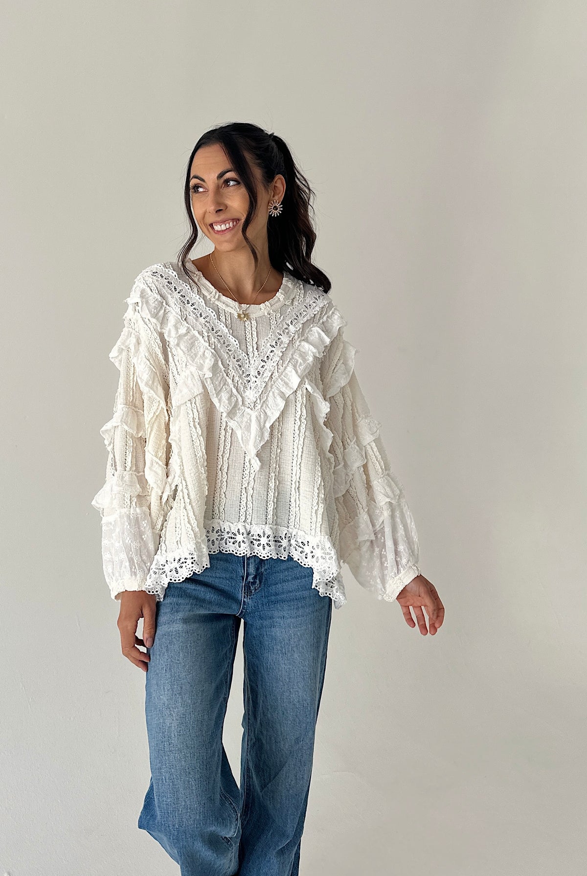 A woman modeling a cream-colored, long-sleeved bohemian-style blouse with intricate lace and ruffle details, paired with blue jeans, against a plain light gray background.