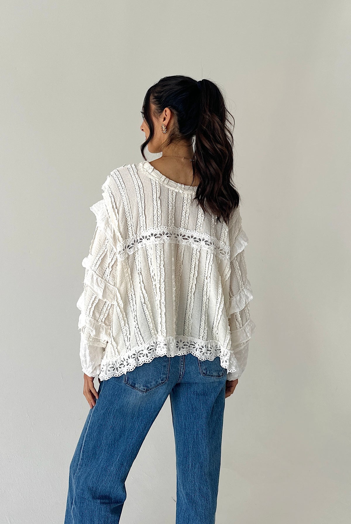 A woman wearing a white long-sleeve blouse with ruffles and lace trim, paired with blue jeans, photographed from behind against a plain light gray studio background.