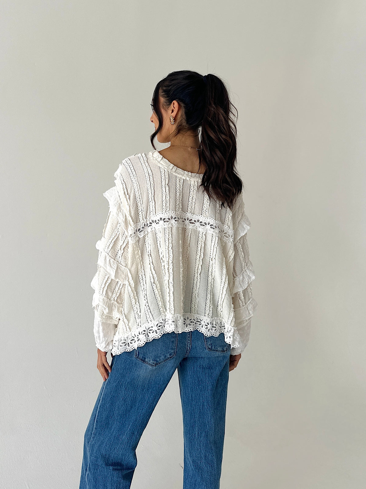 A woman wearing a white long-sleeve blouse with ruffles and lace trim, paired with blue jeans, photographed from behind against a plain light gray studio background.