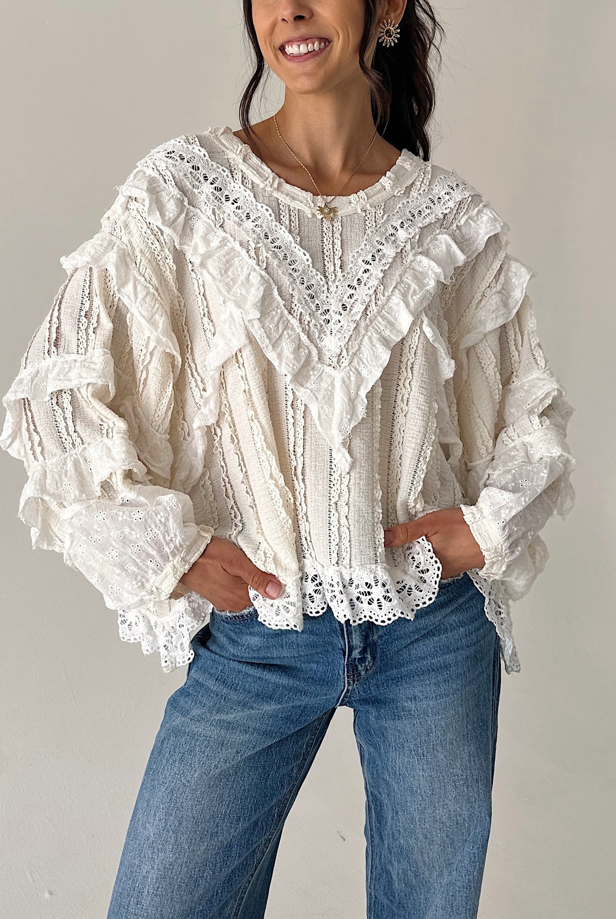 A woman wearing a cream-colored, heavily ruffled and lace-trimmed long-sleeve blouse paired with blue jeans, standing against a plain light gray wall.