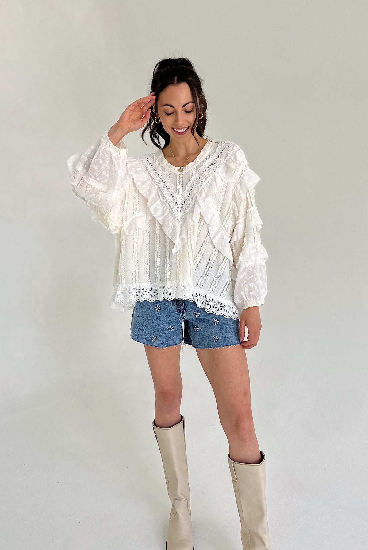 Woman wearing a white lace blouse, denim shorts, and beige knee-high boots against a plain background