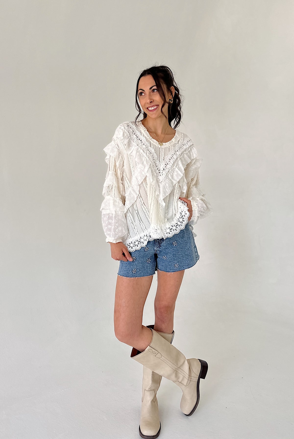 Woman wearing a white lace blouse, denim shorts, and beige knee-high boots against a plain background