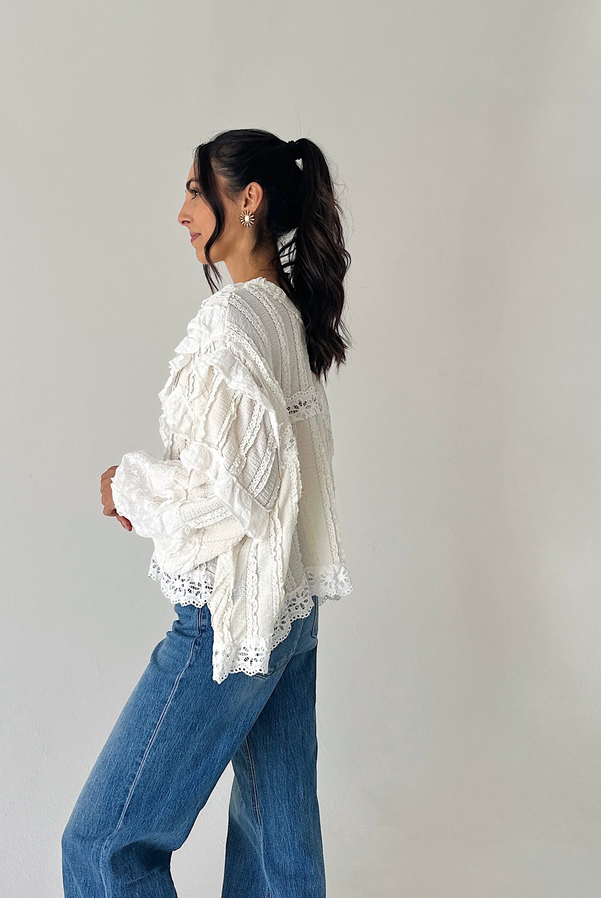 A woman wearing a white lace and crochet long-sleeve blouse with blue jeans, shown from a side profile against a plain light gray studio background.