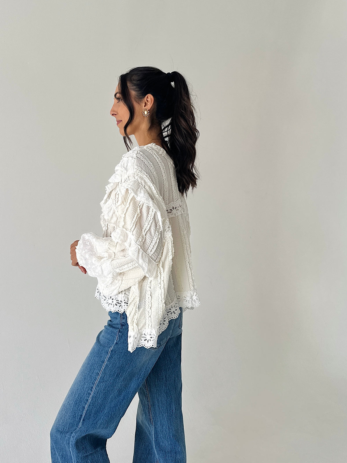 A woman wearing a white lace and crochet long-sleeve blouse with blue jeans, shown from a side profile against a plain light gray studio background.