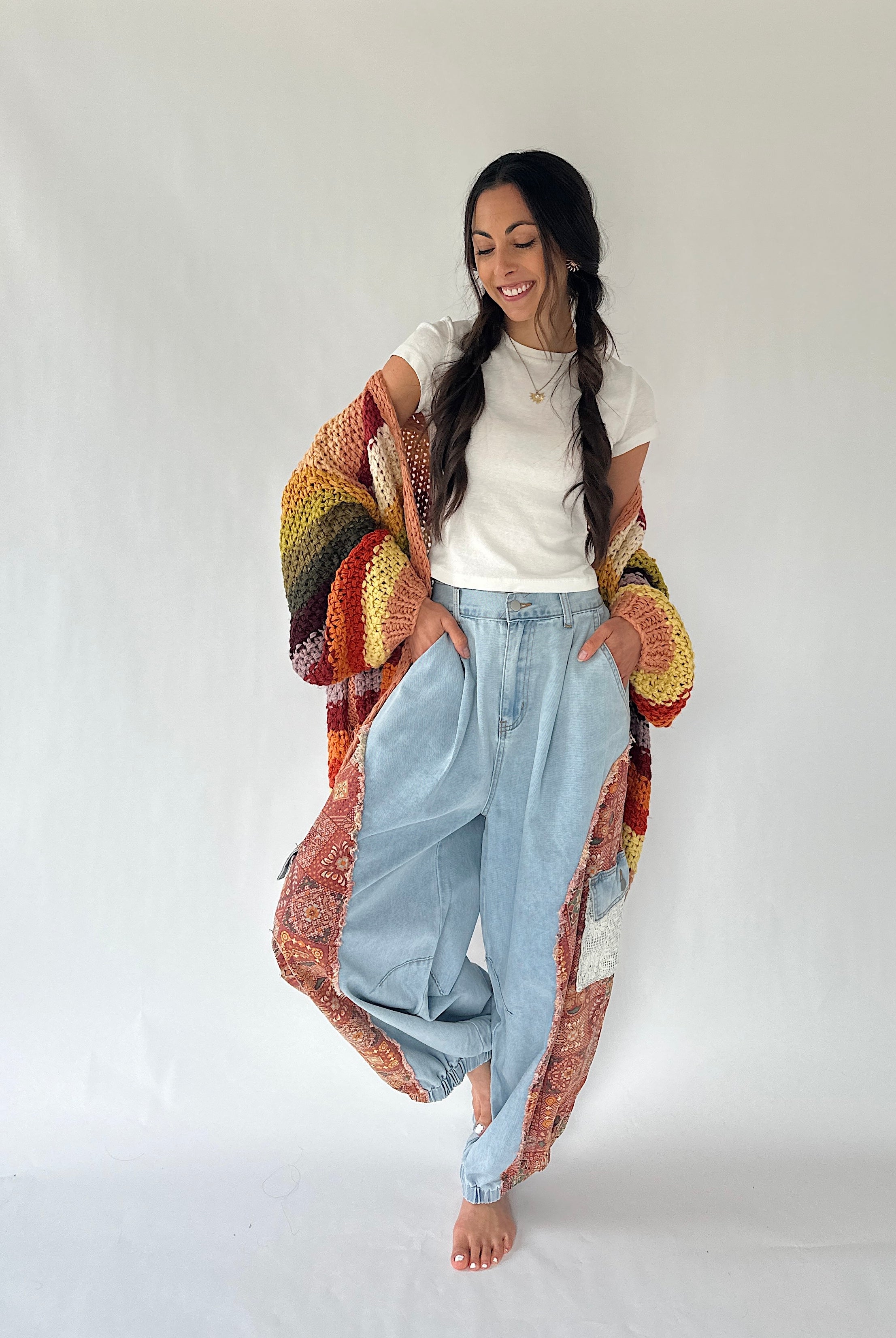A woman modeling a colorful patchwork knit cardigan over a white t-shirt and light-wash wide-leg jeans, standing against a plain white studio background.