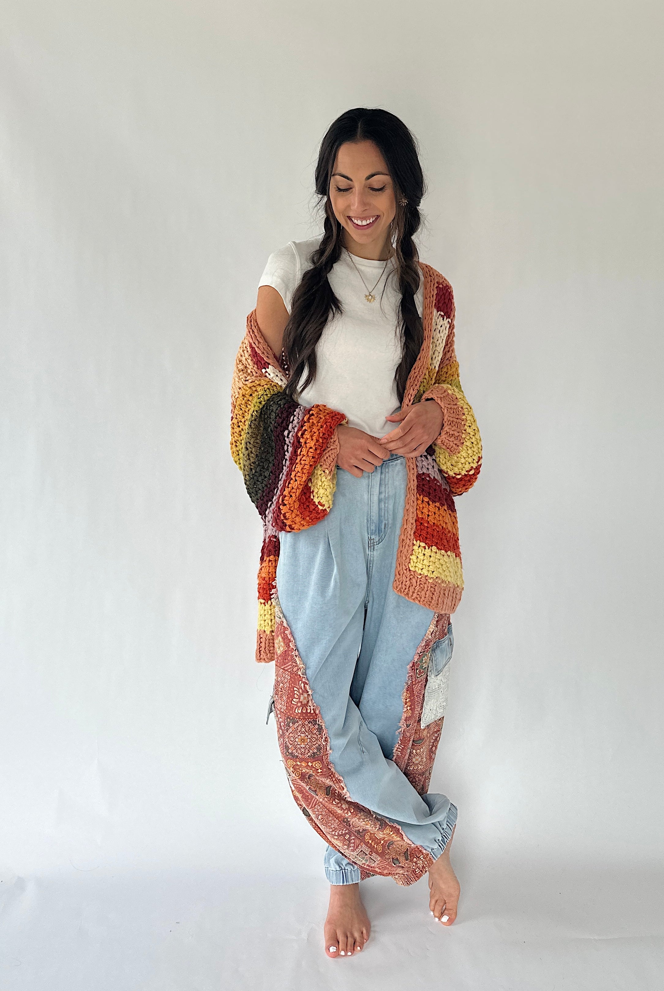 A woman stands barefoot against a plain white studio background, wearing a long, loose, patchwork-style cardigan or cover-up with a multicolor crochet and paisley pattern in orange, yellow, red, brown, and cream over a white top and light blue jeans.